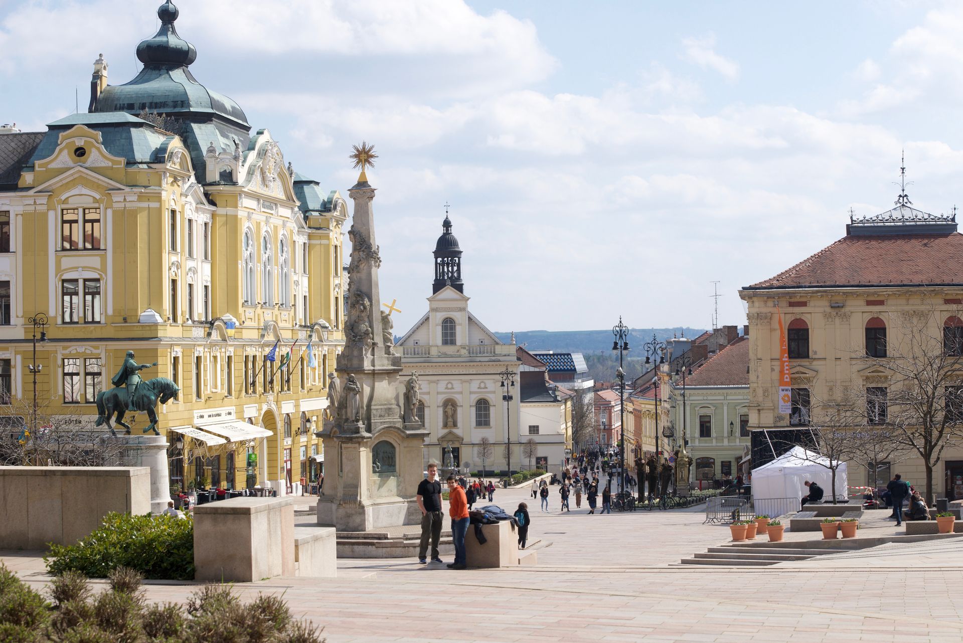 A square with ornate buildings, a column, and people walking; sunny day in a European city.