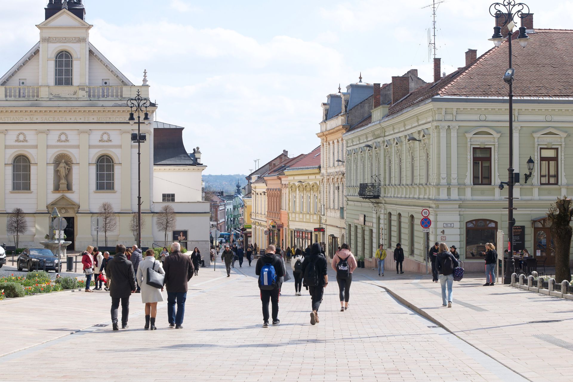 People walking on a cobblestone street lined with buildings, a church, and shops. Sunny day.