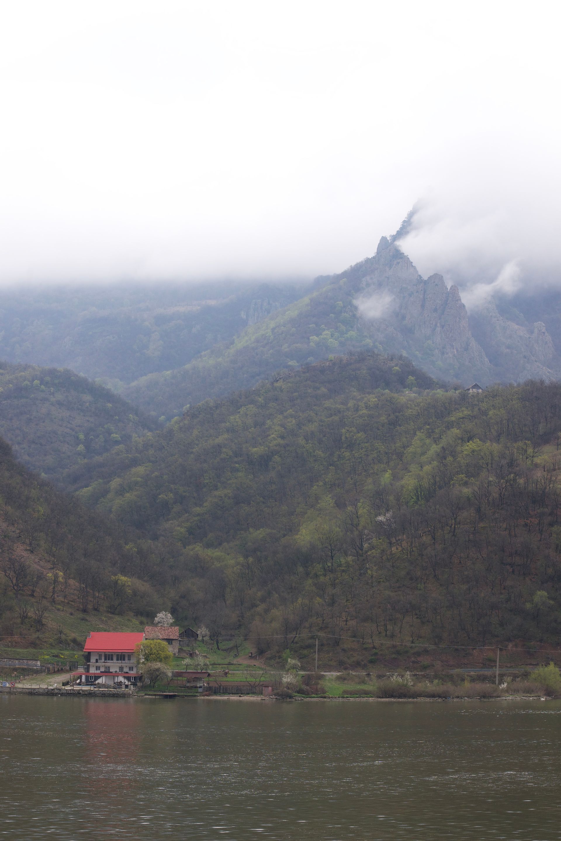 Red-roofed building by water with mountains in the background, obscured by mist and overcast sky.