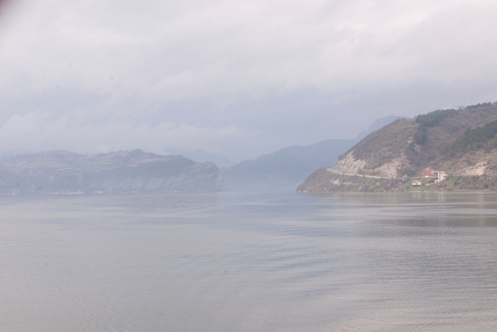 Calm water reflecting mountains under a hazy, overcast sky.