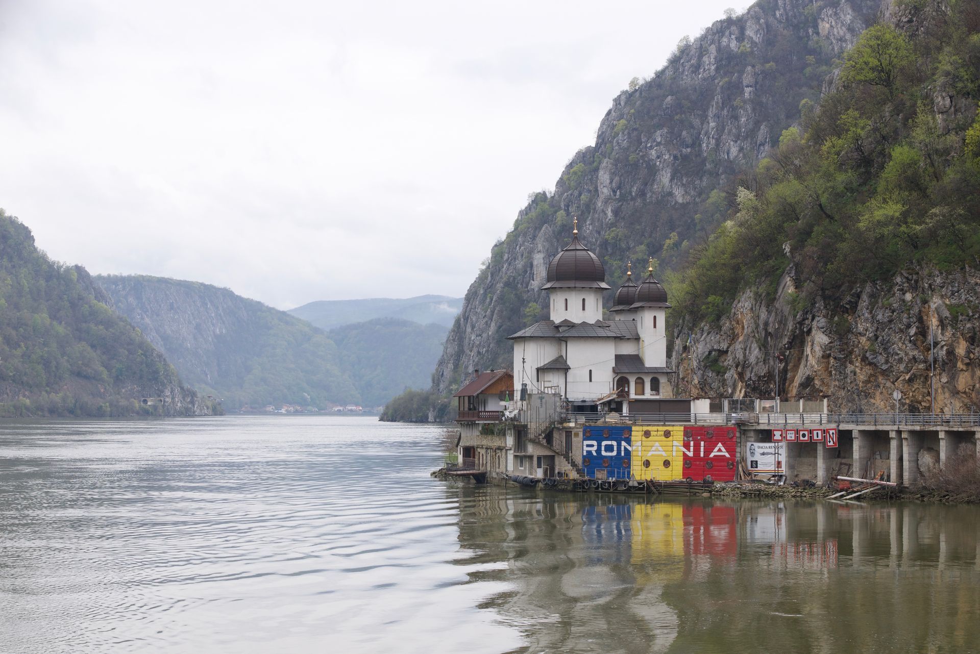 Monastery on a river, with colorful buildings against a cliff, and mountains in the background.