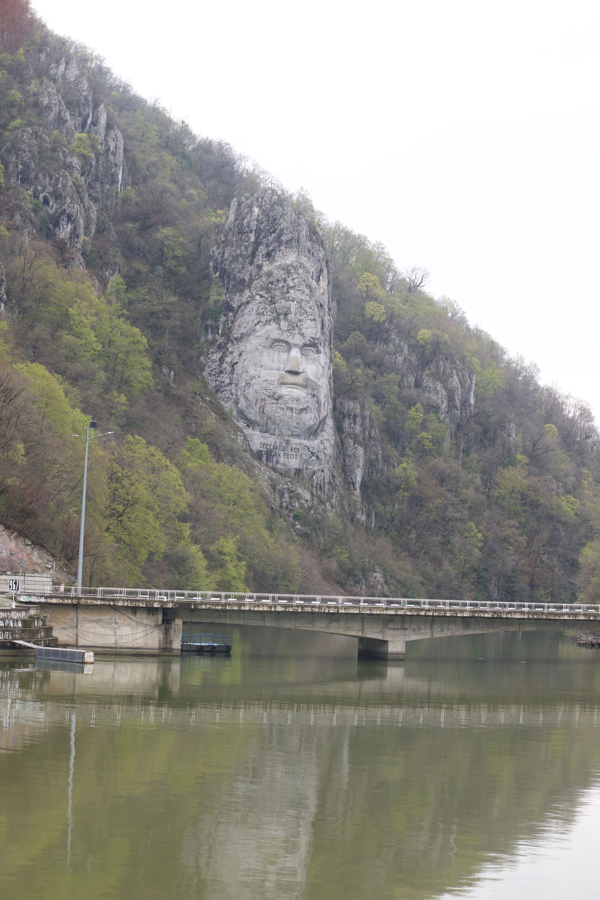 Giant rock carving of a face on a cliffside, overlooking a river with a bridge, cars, and trees.
