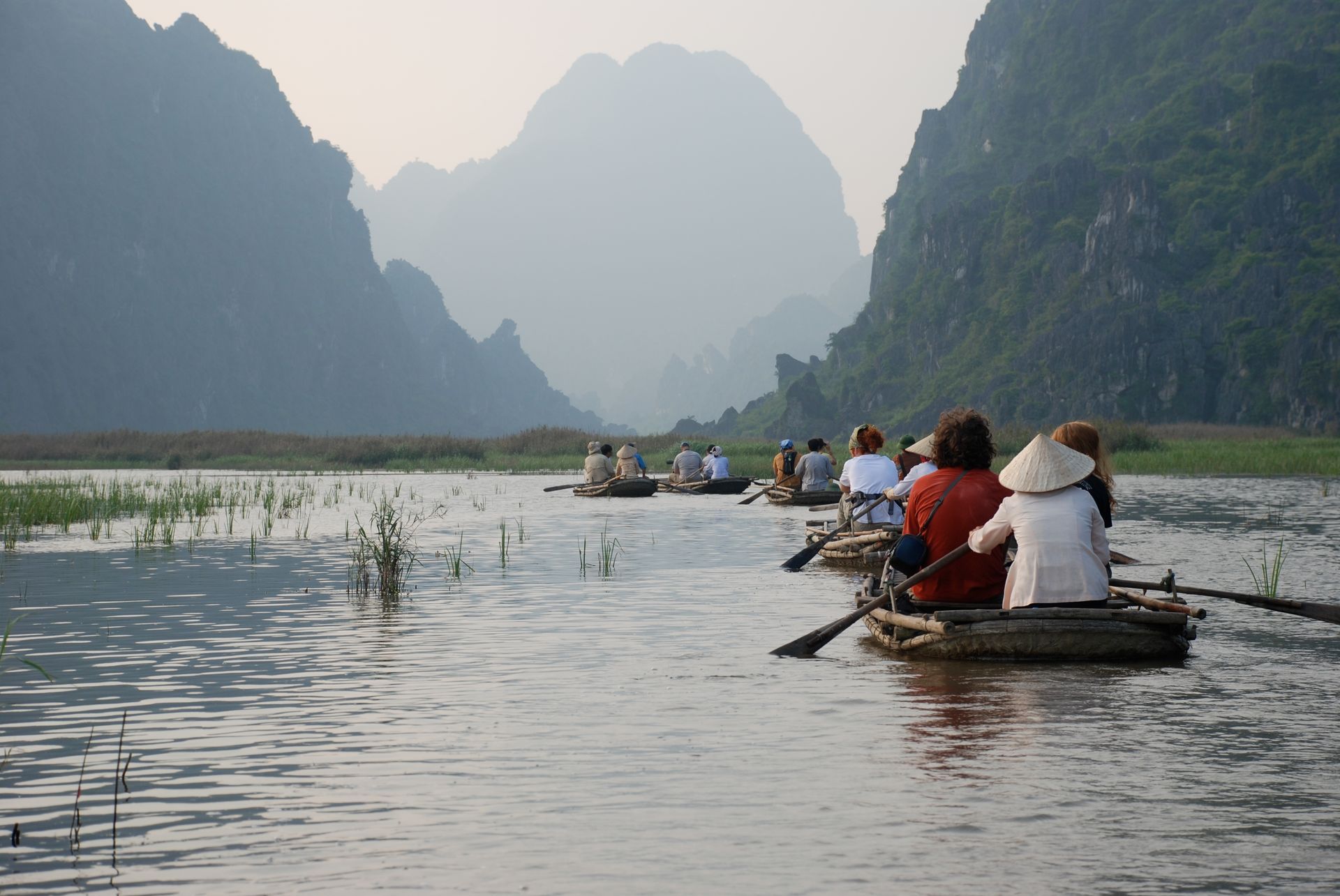 Rowboats on a river, silhouetted mountains in background. People in hats and shirts enjoy a scenic ride.