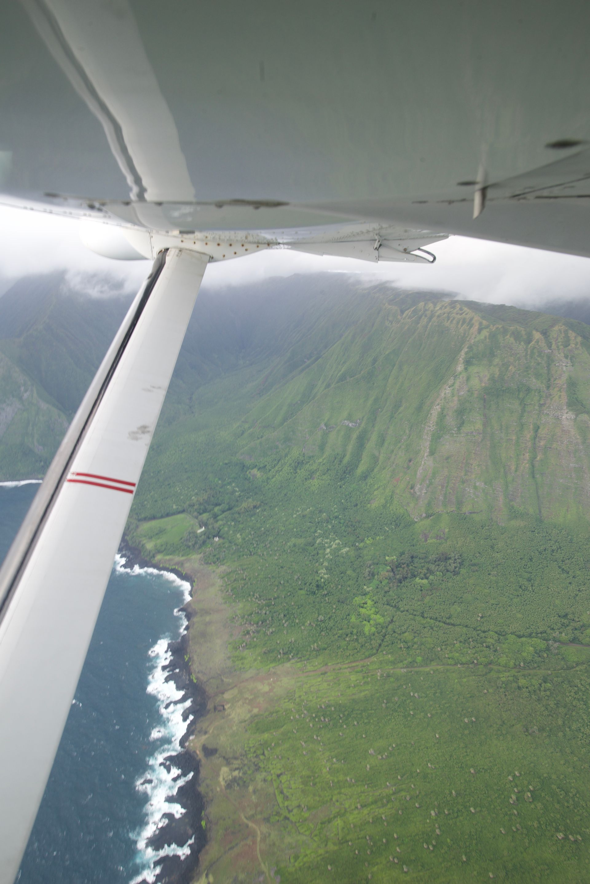 View from a small plane overlooking a lush green coastline and mountain. Cloudy sky and ocean visible.