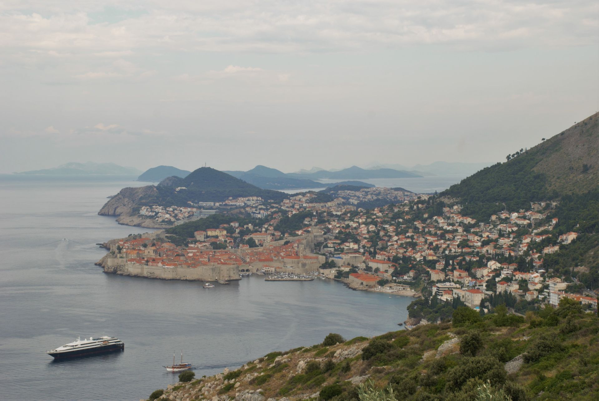 Coastal view of Dubrovnik, Croatia, with walled city, sea, mountains, and boat under overcast sky.