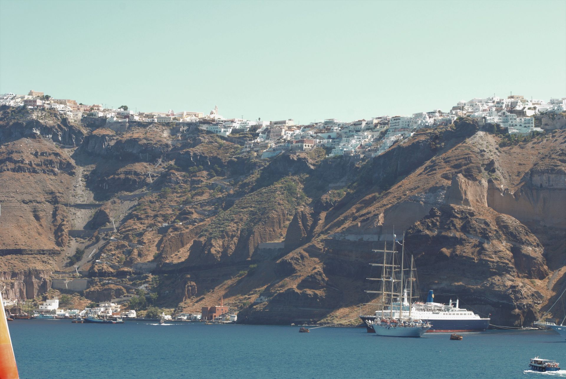 Sailboat in Aegean Sea with white buildings perched on a cliffside in Santorini, Greece.