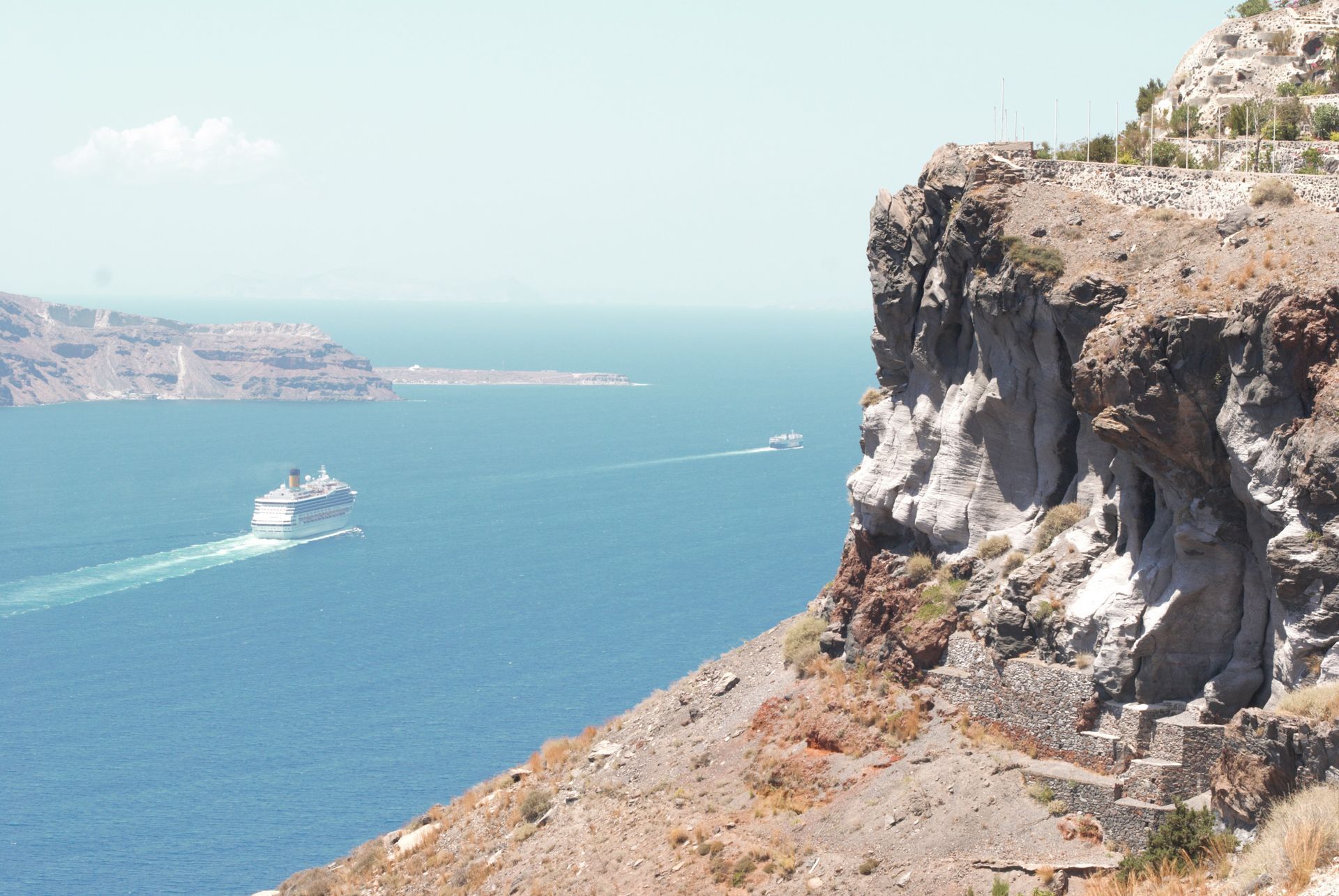 Cliff overlooking a blue sea with a cruise ship and island in the distance.