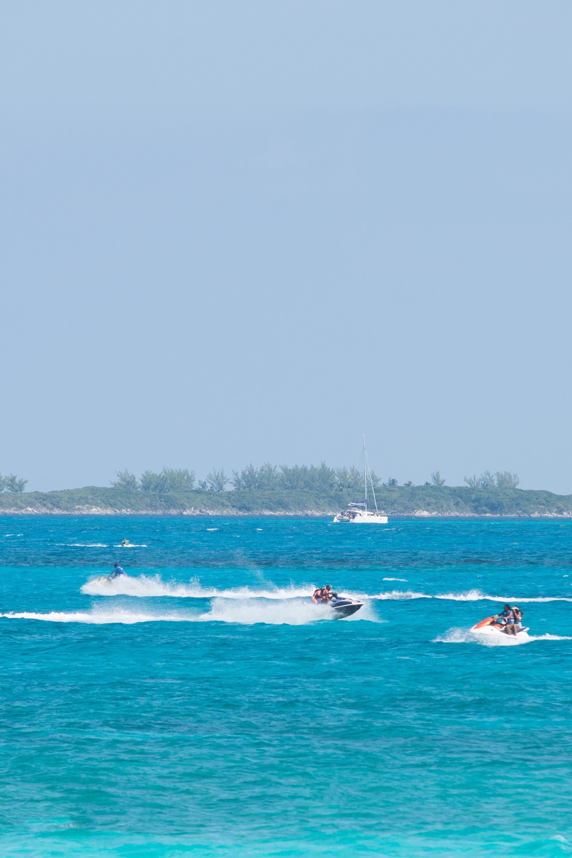 Jet skis speeding on turquoise ocean, with a distant island and sailboat.