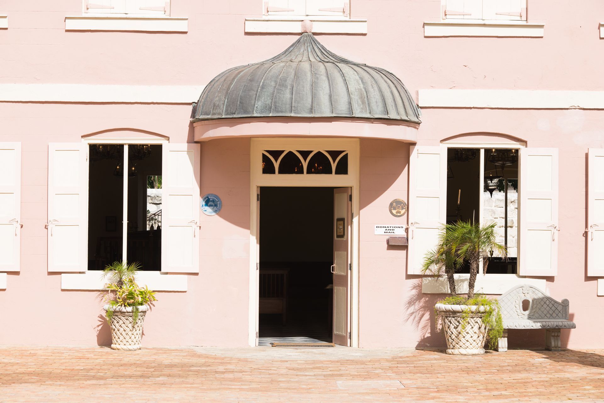 Pink building with dome-topped entrance, white-framed windows, and potted plants outside.