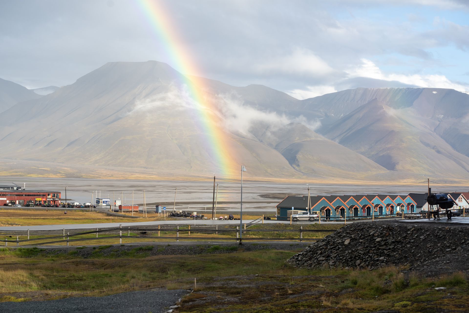 A rainbow is visible over a small town with mountains in the background in Svalbard.