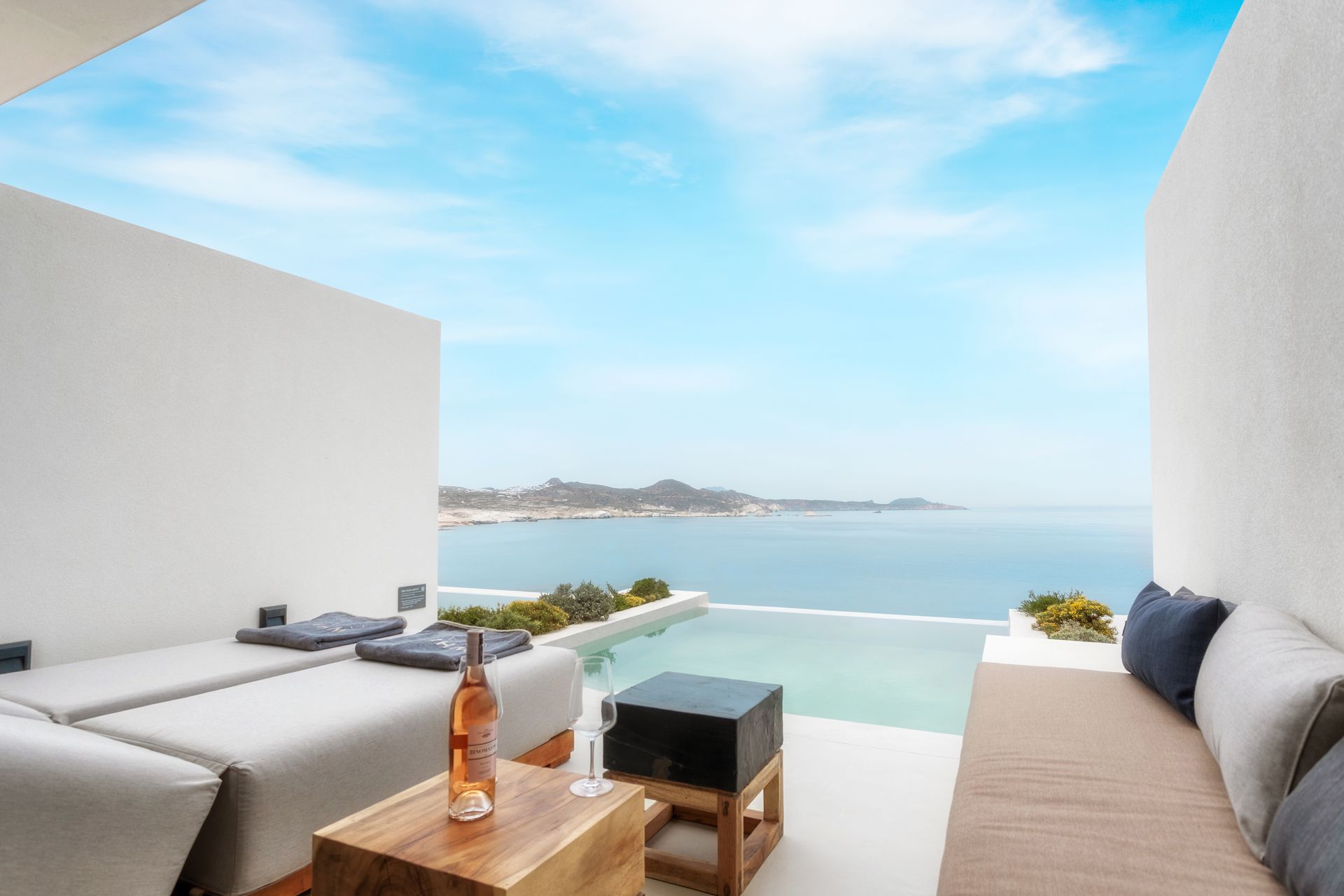 A bottle of wine is on a table in front of a swimming pool overlooking the ocean at Domes White Coast Hotel in Milos, Greece.