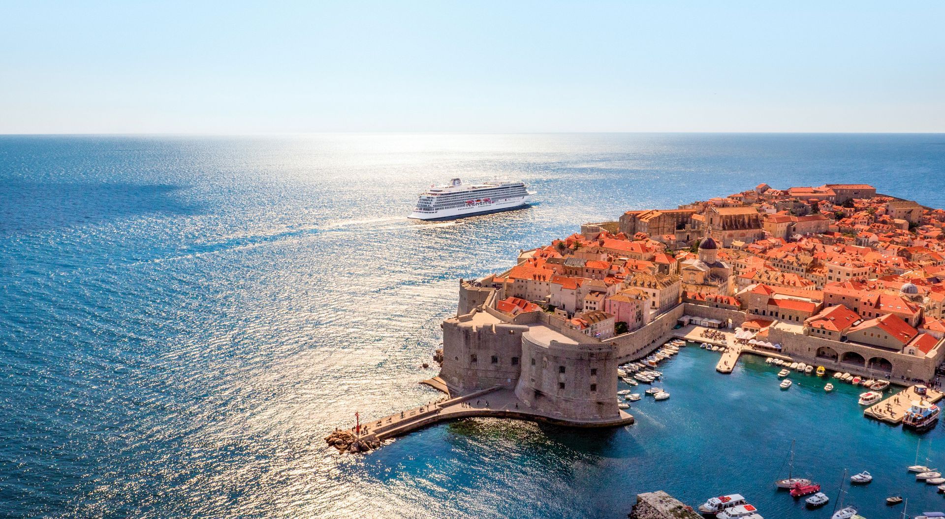 Coastal scene: Cruise ship near historic orange-roofed city walls, docked harbor, blue sea, bright sunshine.