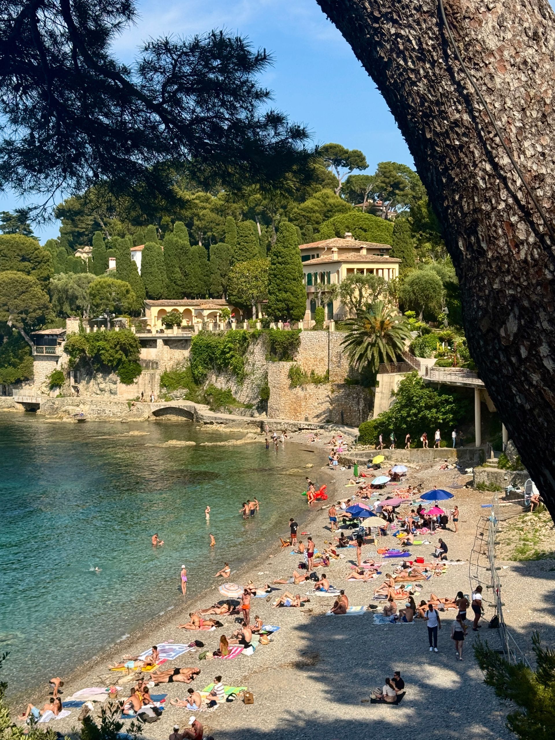 Beach with people sunbathing; cliffs, buildings, and clear water under a blue sky.