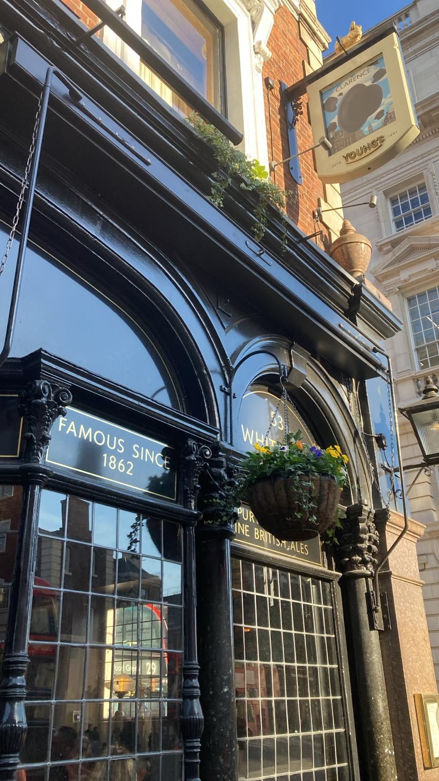 Black pub facade with hanging flower basket. Sign reads 