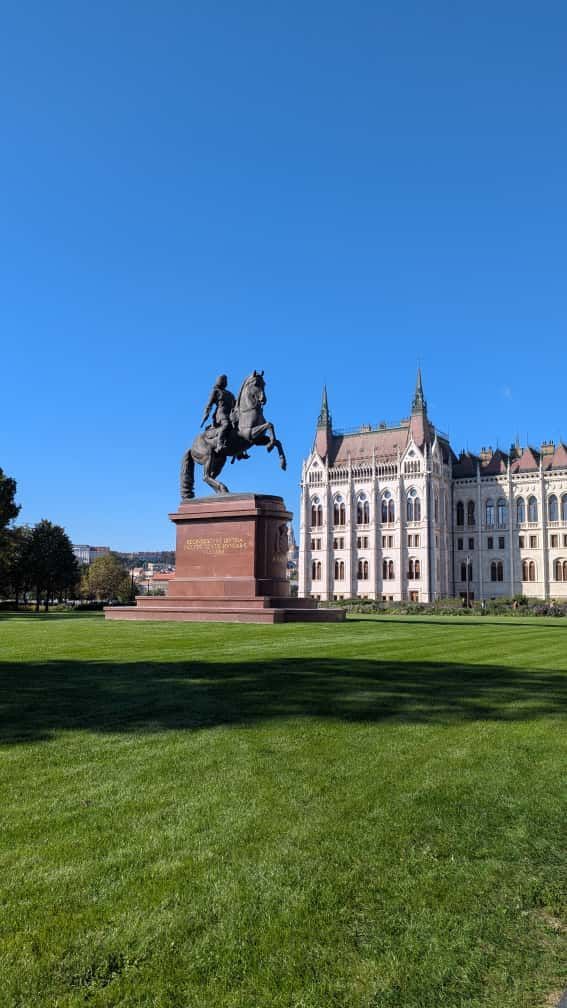 Bronze equestrian statue in front of a white building with towers, on a grassy lawn under a blue sky.