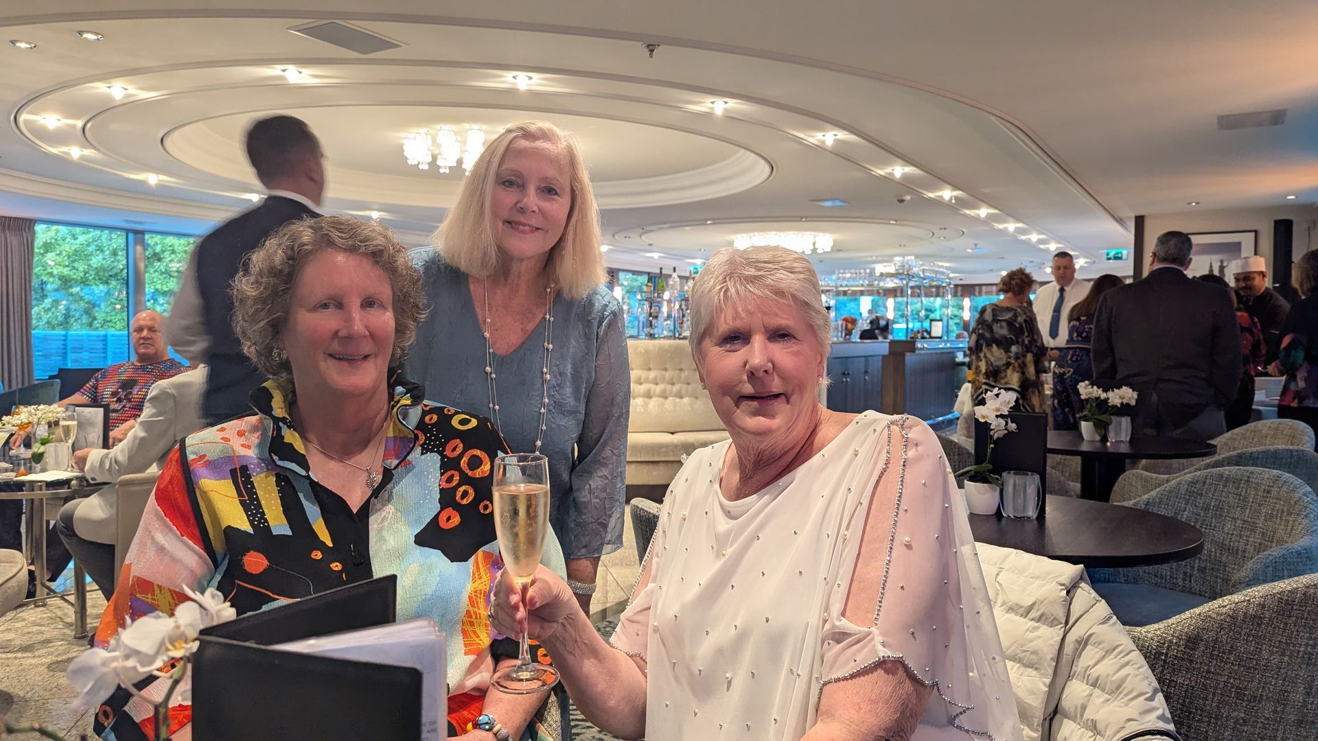 Three women raise champagne glasses in a lounge, smiling. Others gather in the background, near a window.