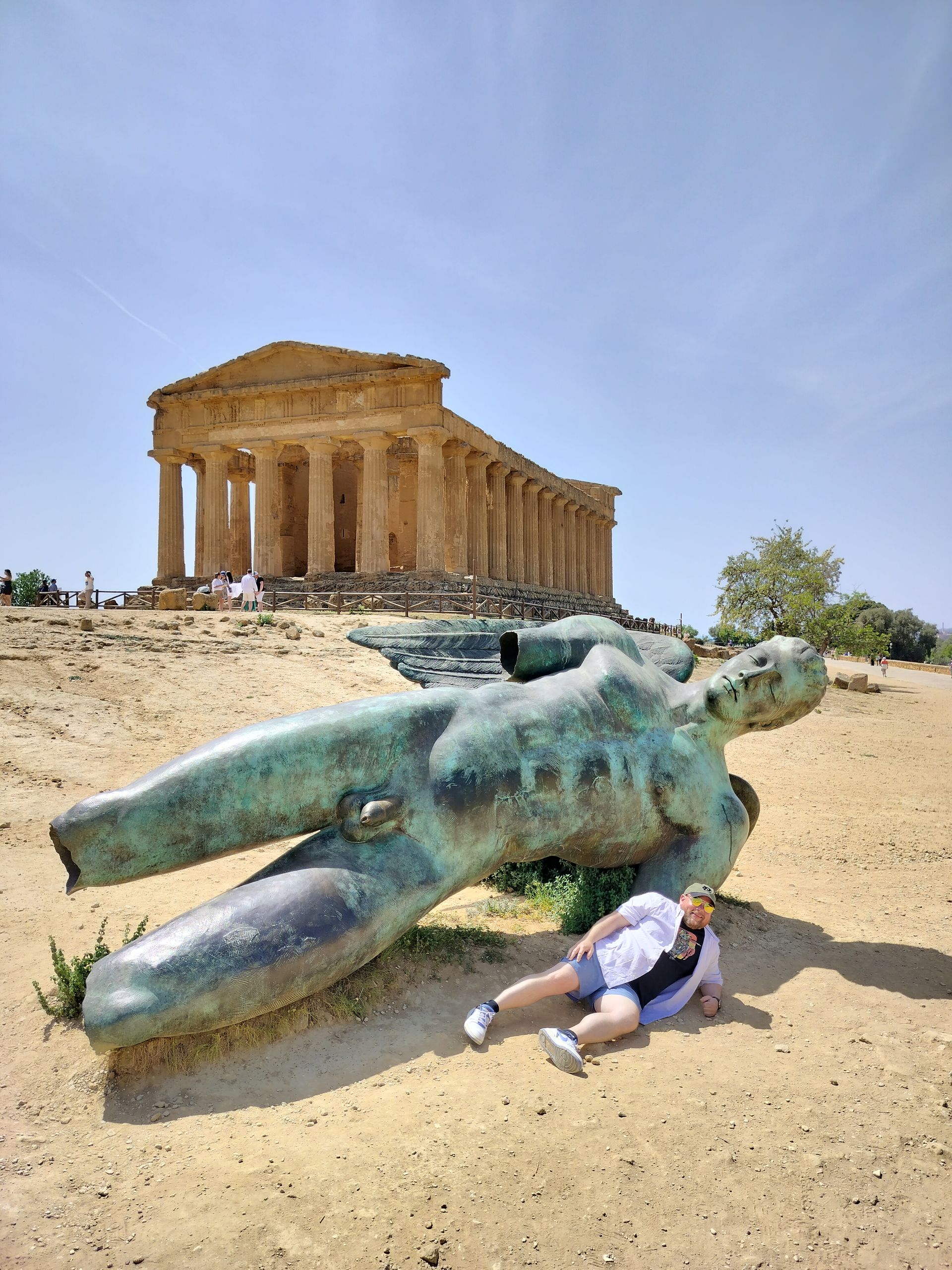 Person reclining near a bronze statue, ancient temple in background, sunny day.