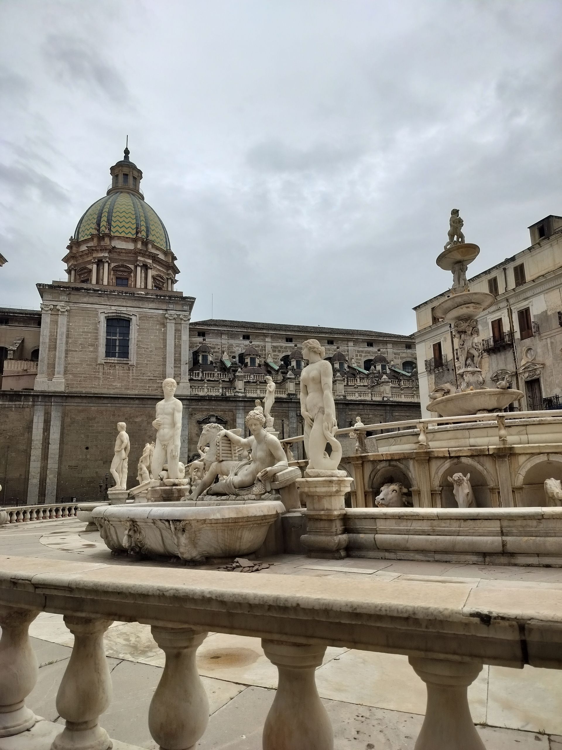 Fountain with statues in a plaza, light stone buildings, green domed church, cloudy sky in Palermo, Italy.