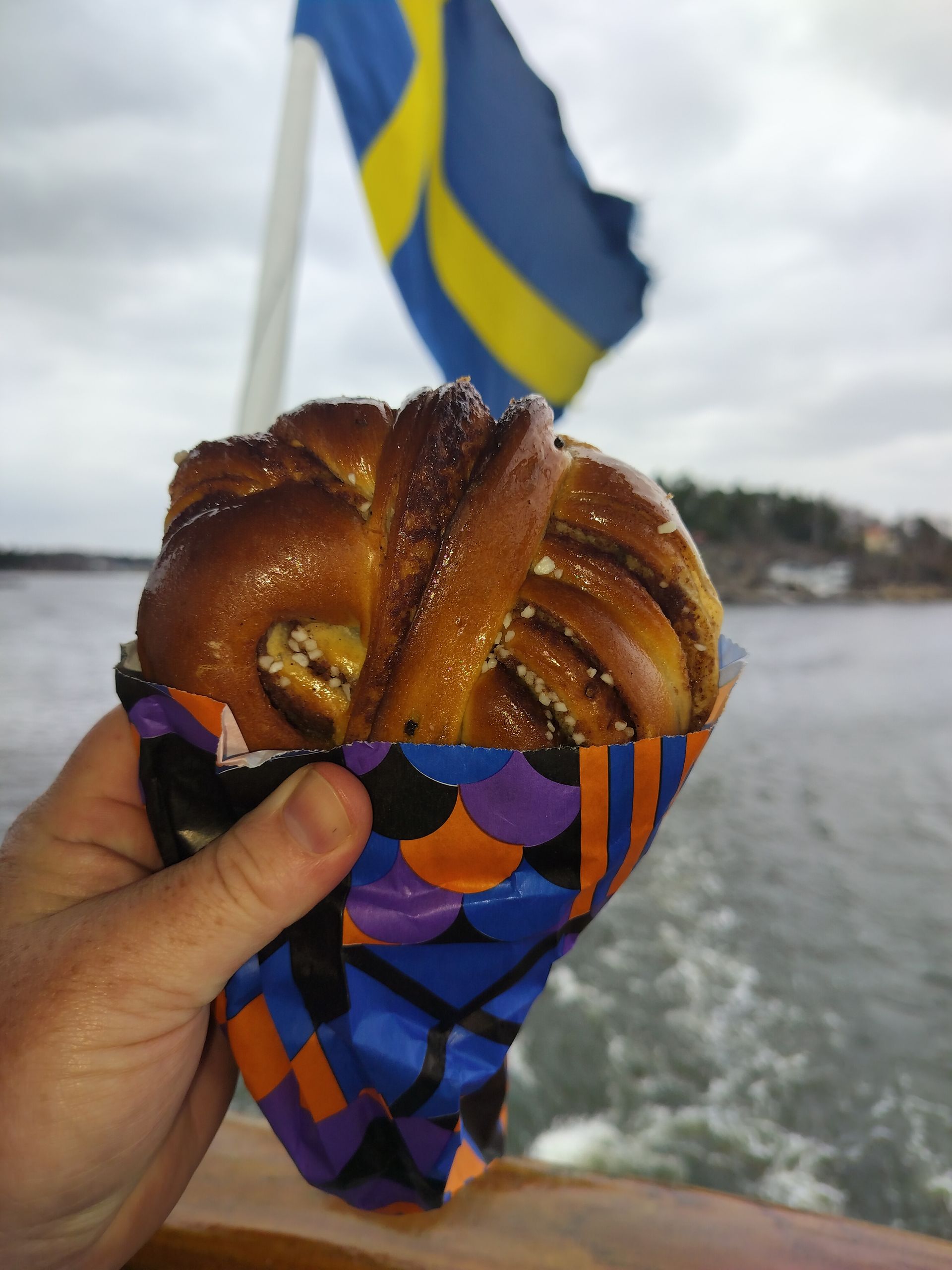 Hand holding a cinnamon bun, Swedish flag in the background on a boat.