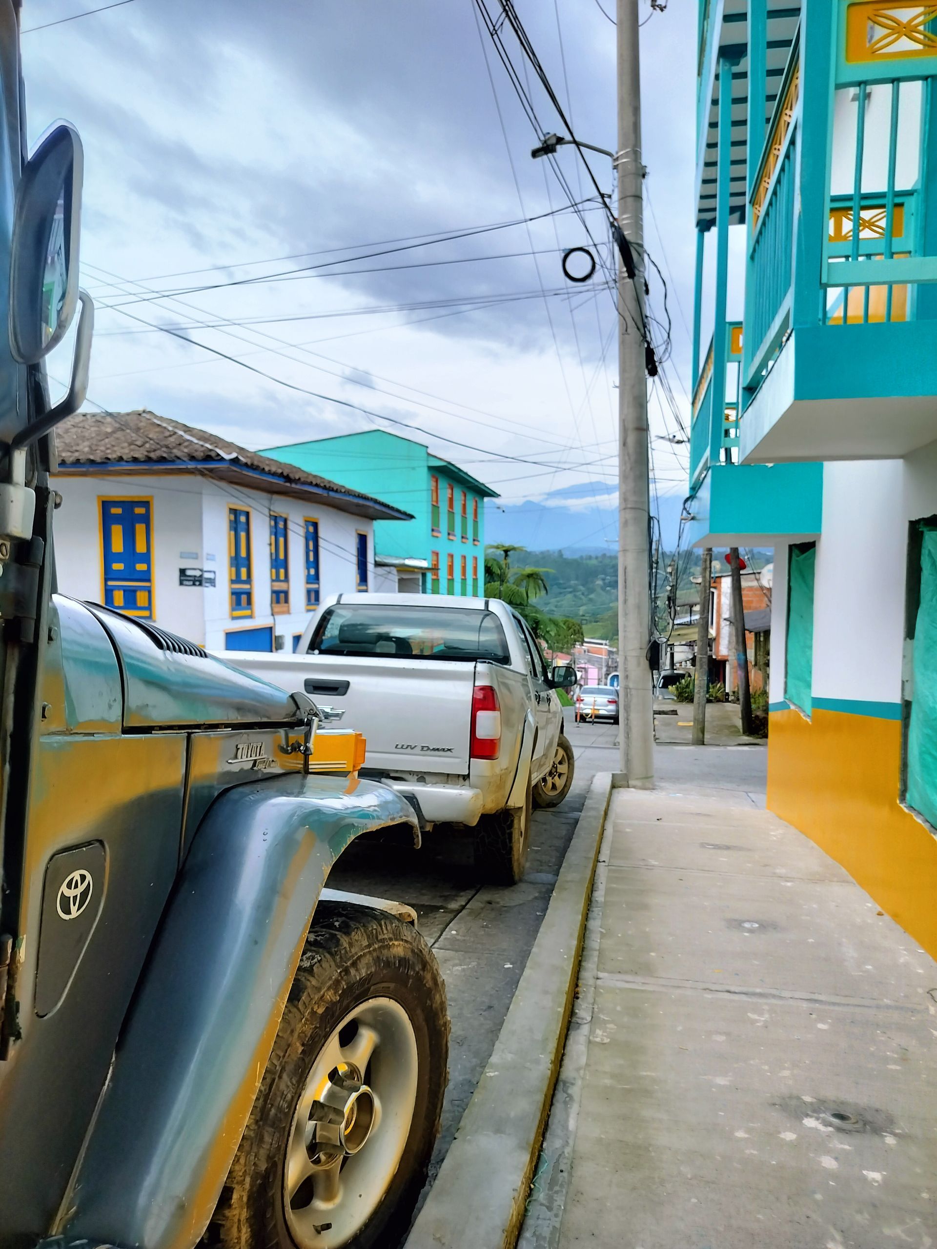 Jeep parked on a narrow street lined with colorful buildings. A white pickup truck is parked in front.