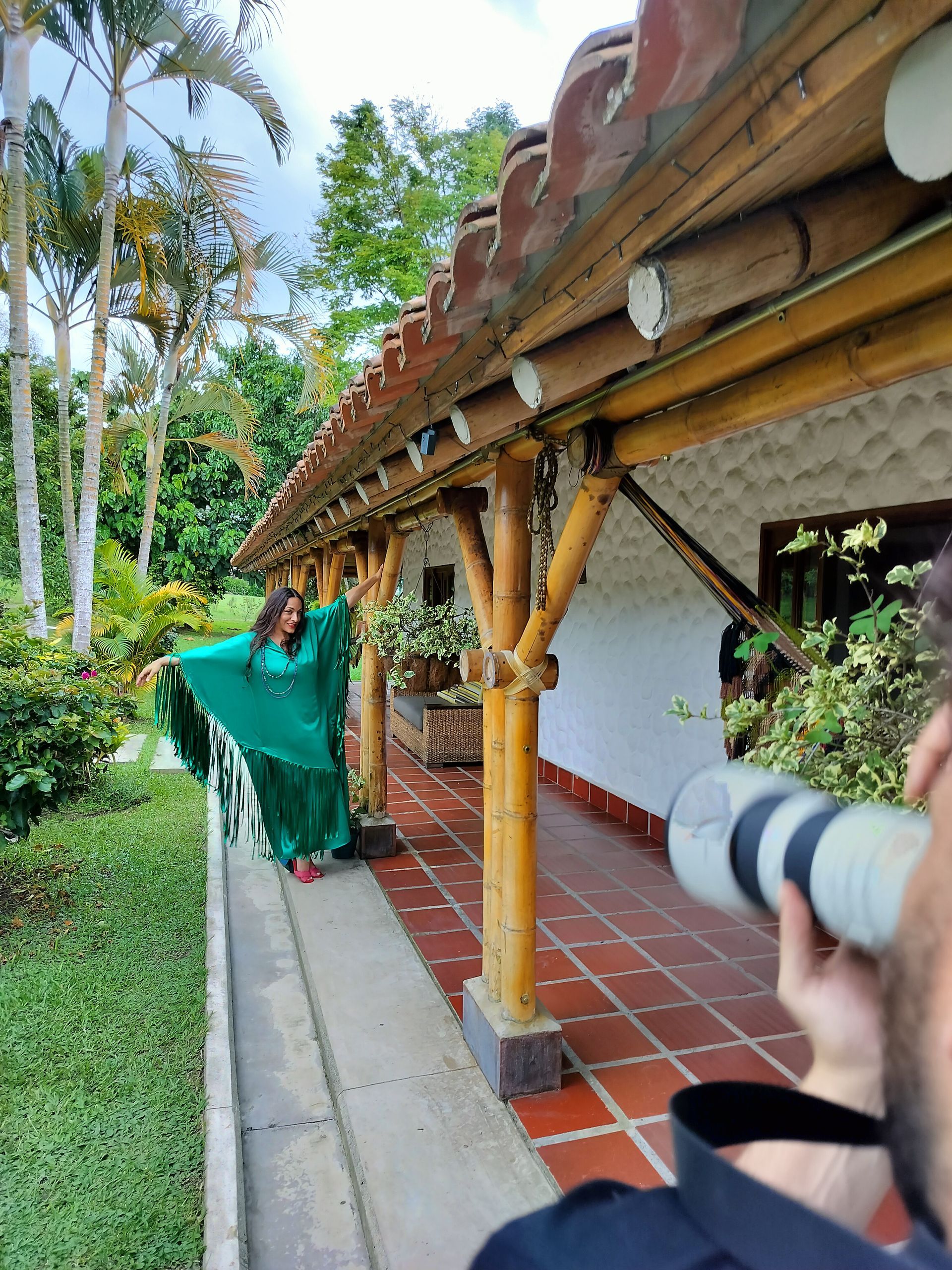 Woman poses in green outfit, photographer captures her on a porch with terracotta tiles and bamboo supports.