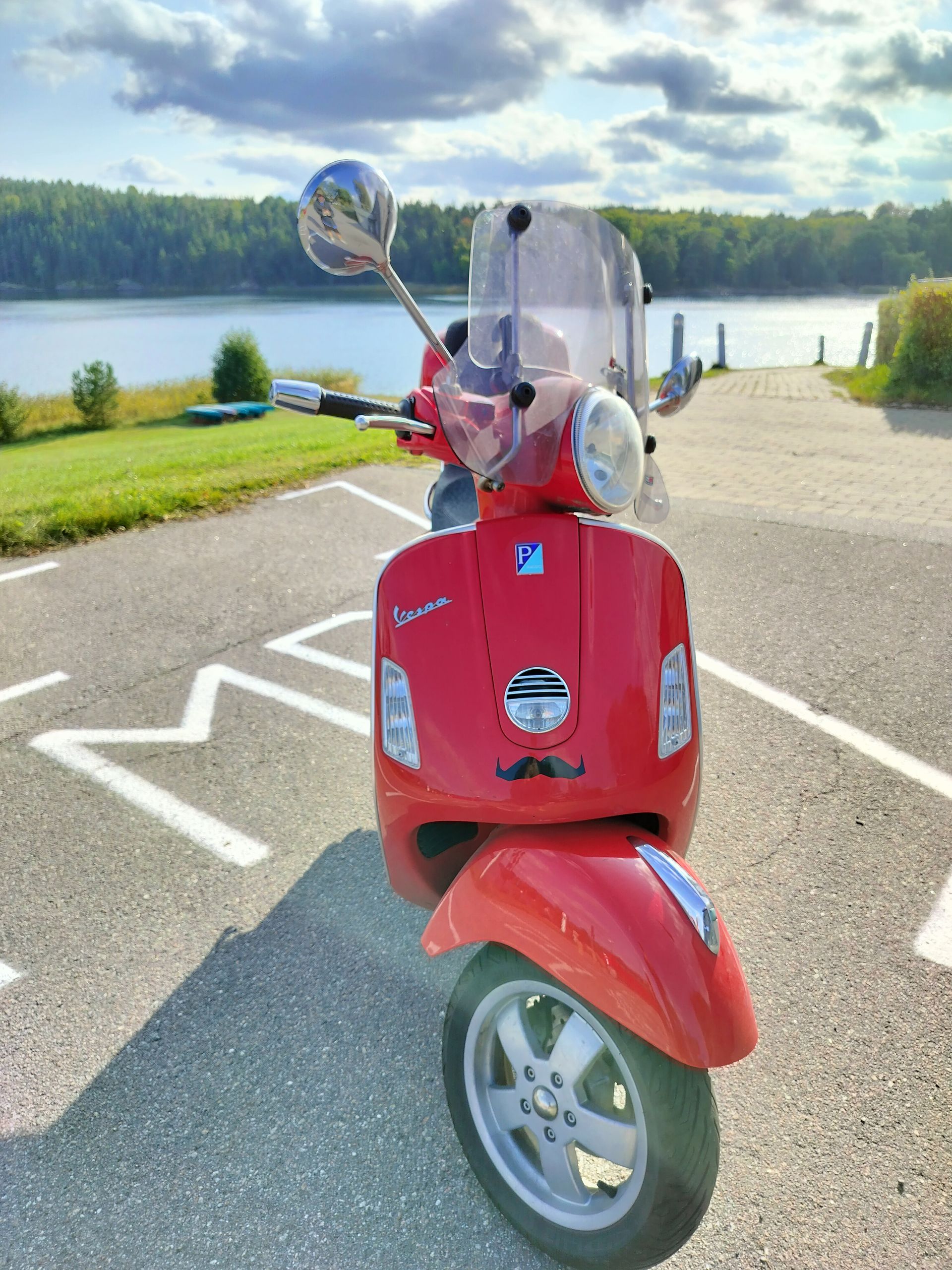 Red Vespa scooter parked on asphalt near a lake and trees.