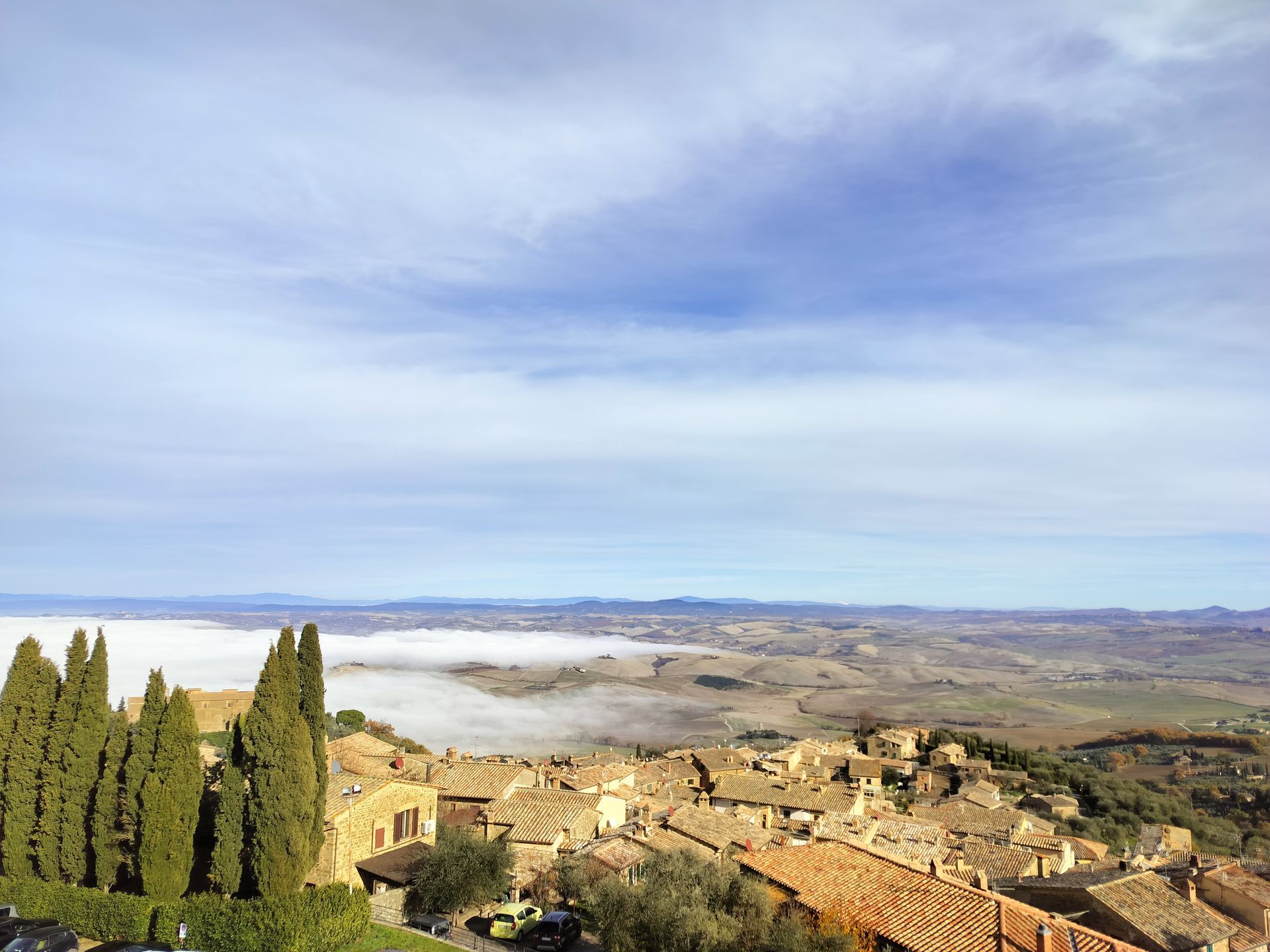 View of a hilltop village with red-tiled roofs, cypress trees, and a valley filled with mist under a blue sky.