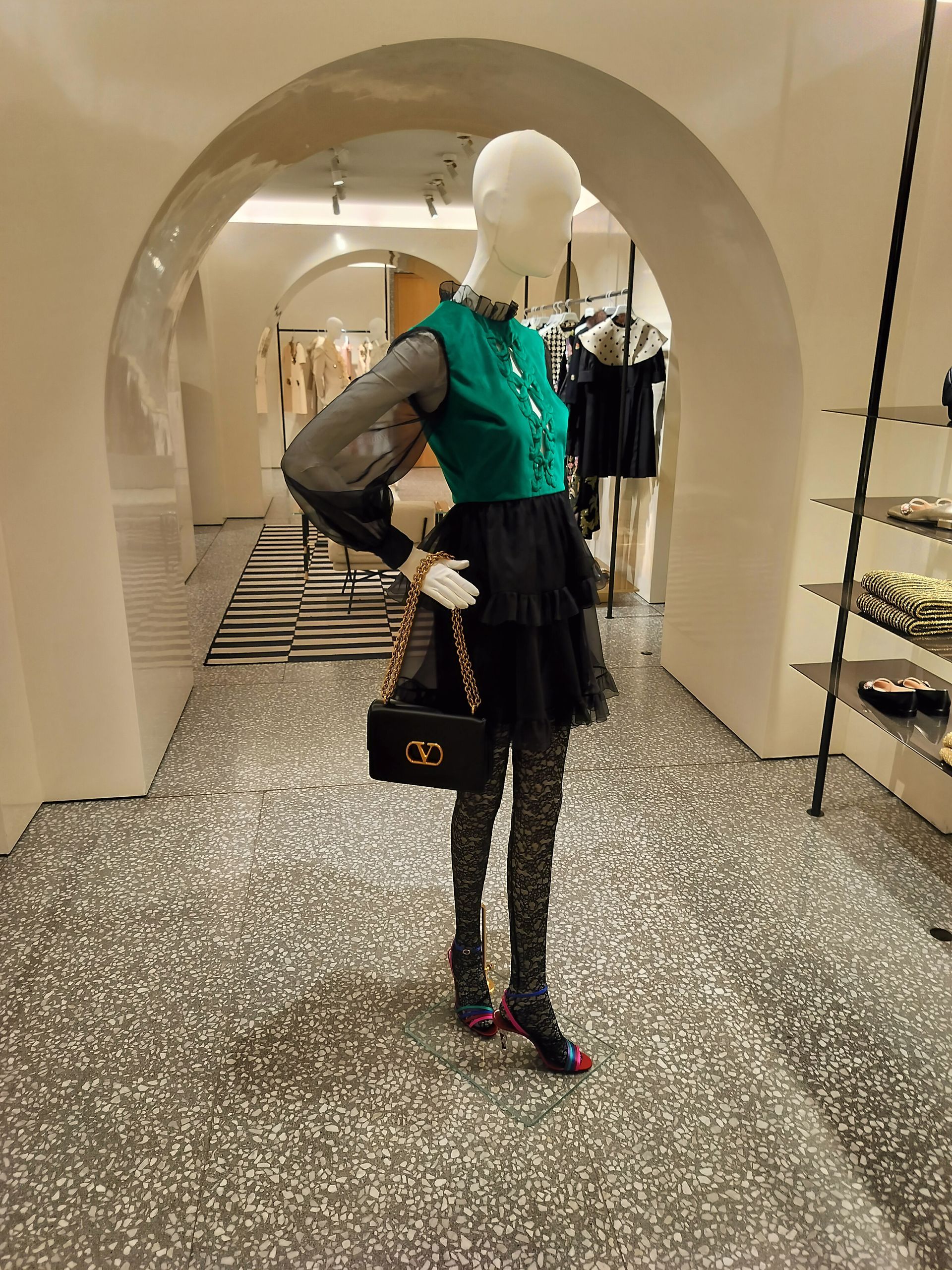 Mannequin in a store wearing a green top, black skirt, and patterned tights, holding a black purse.