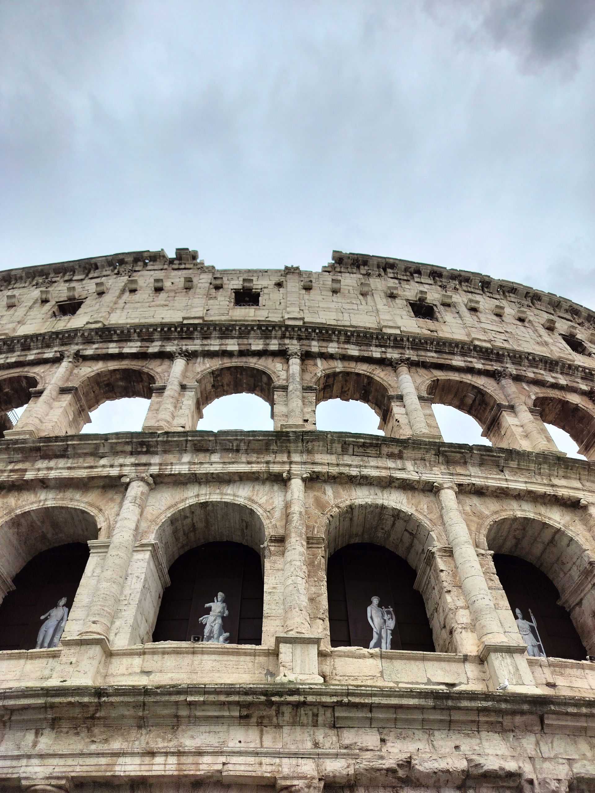 Colosseum in Rome, Italy, with arched windows and statues, against a cloudy sky.