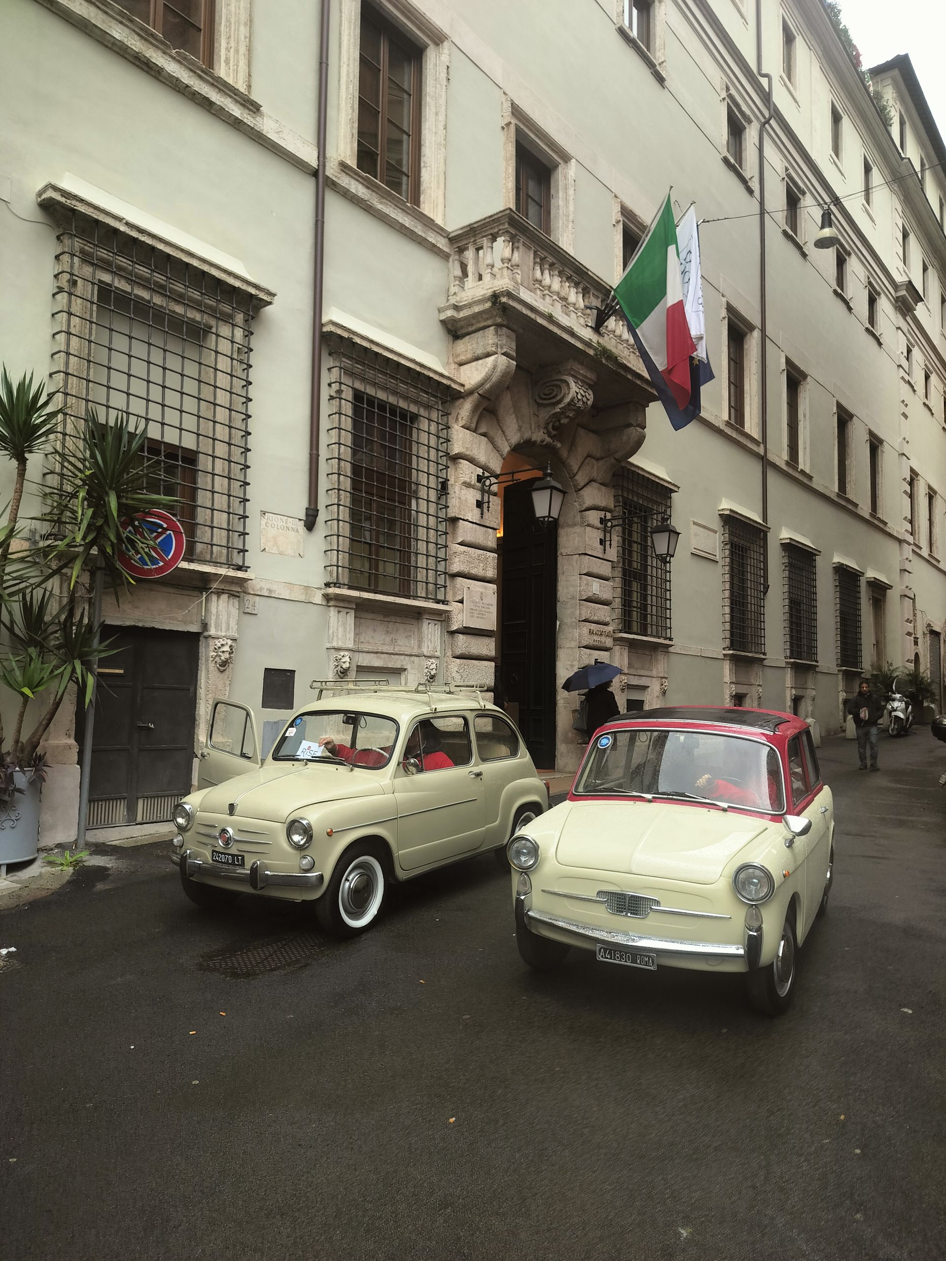 Two vintage Fiat cars parked in front of a light-colored building with an Italian flag.
