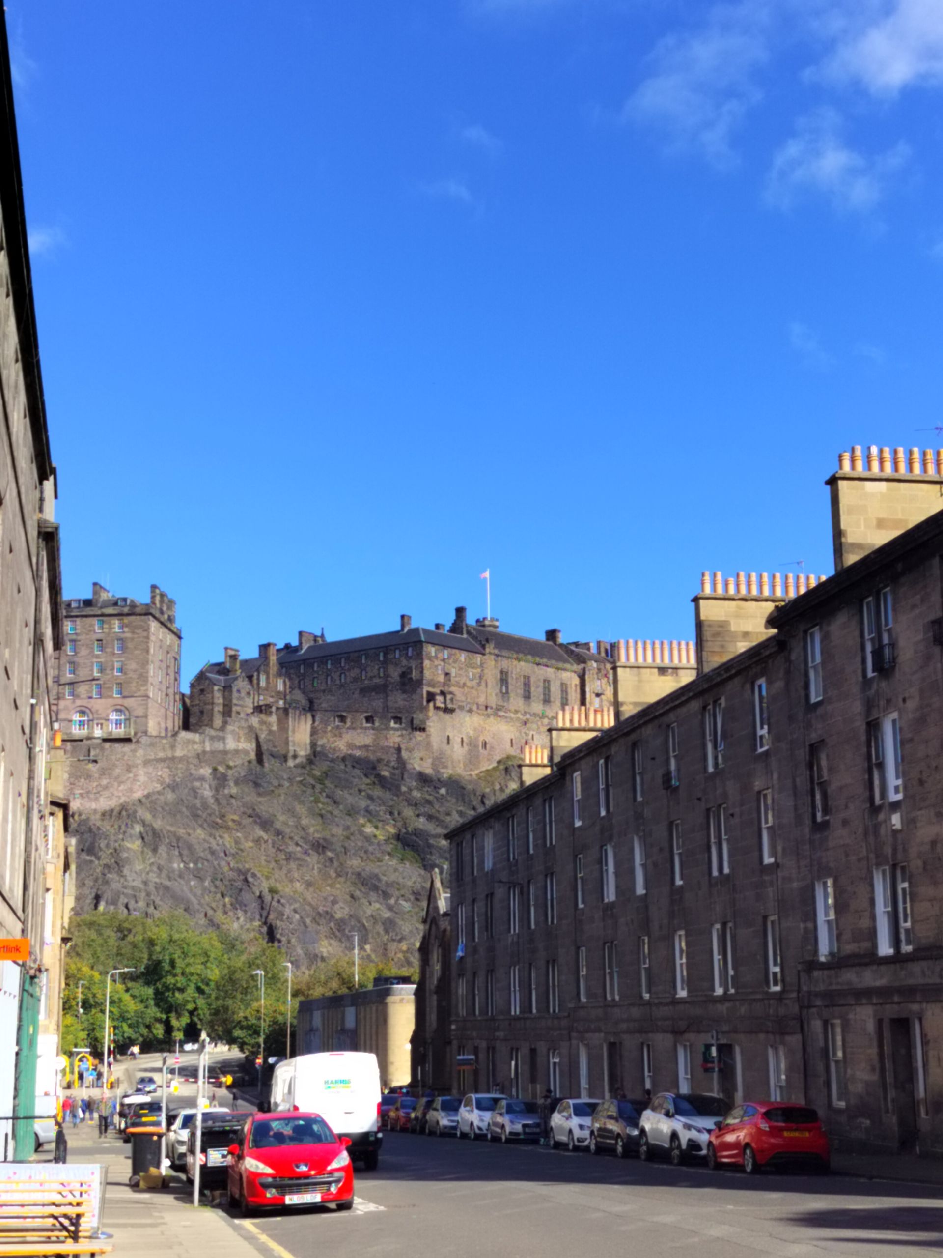 Edinburgh Castle overlooking a street lined with stone buildings and parked cars, under a clear blue sky.