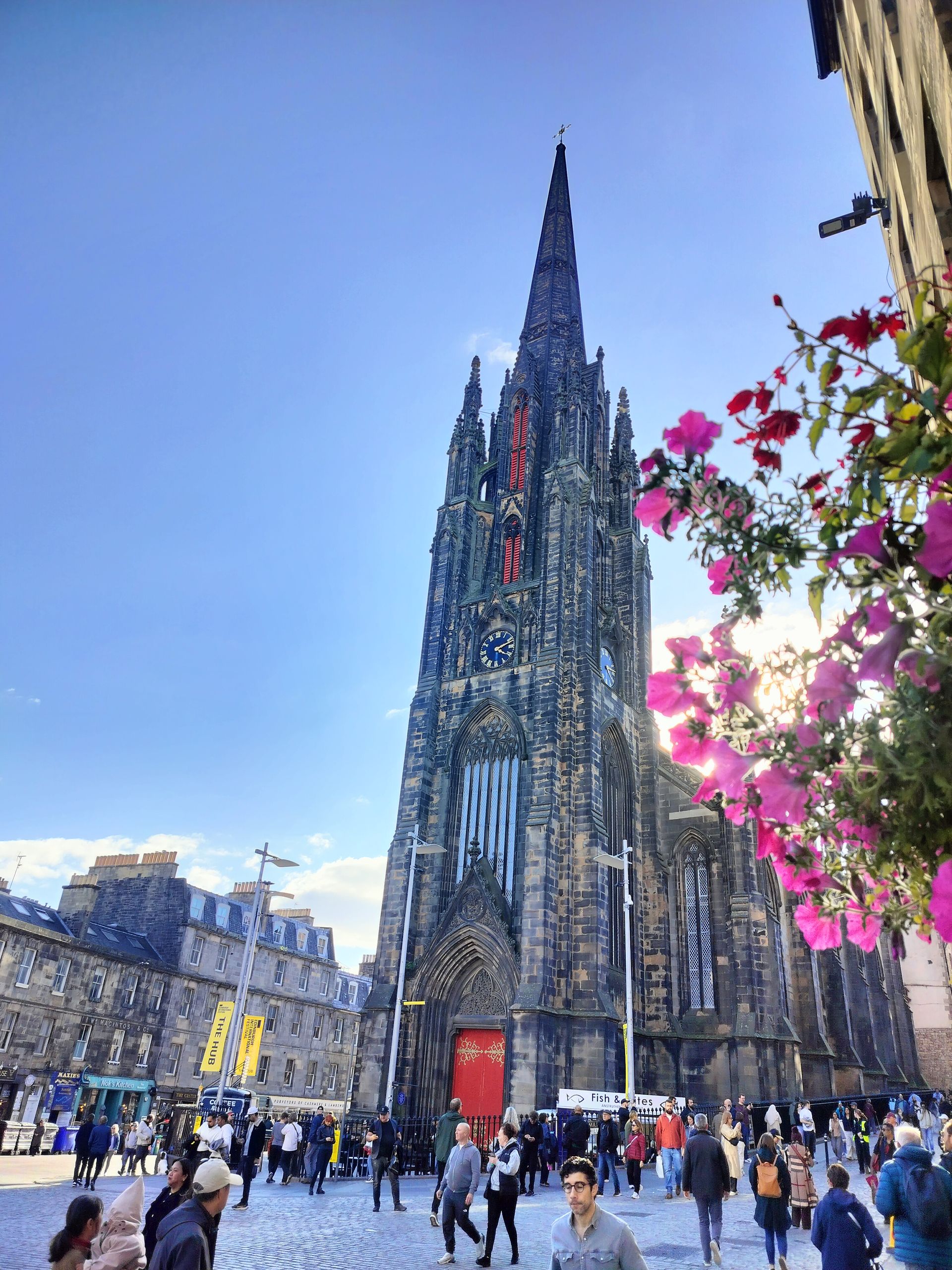 A tall, gothic church with a spire dominates a crowded city square on a sunny day. Pink flowers frame the scene.