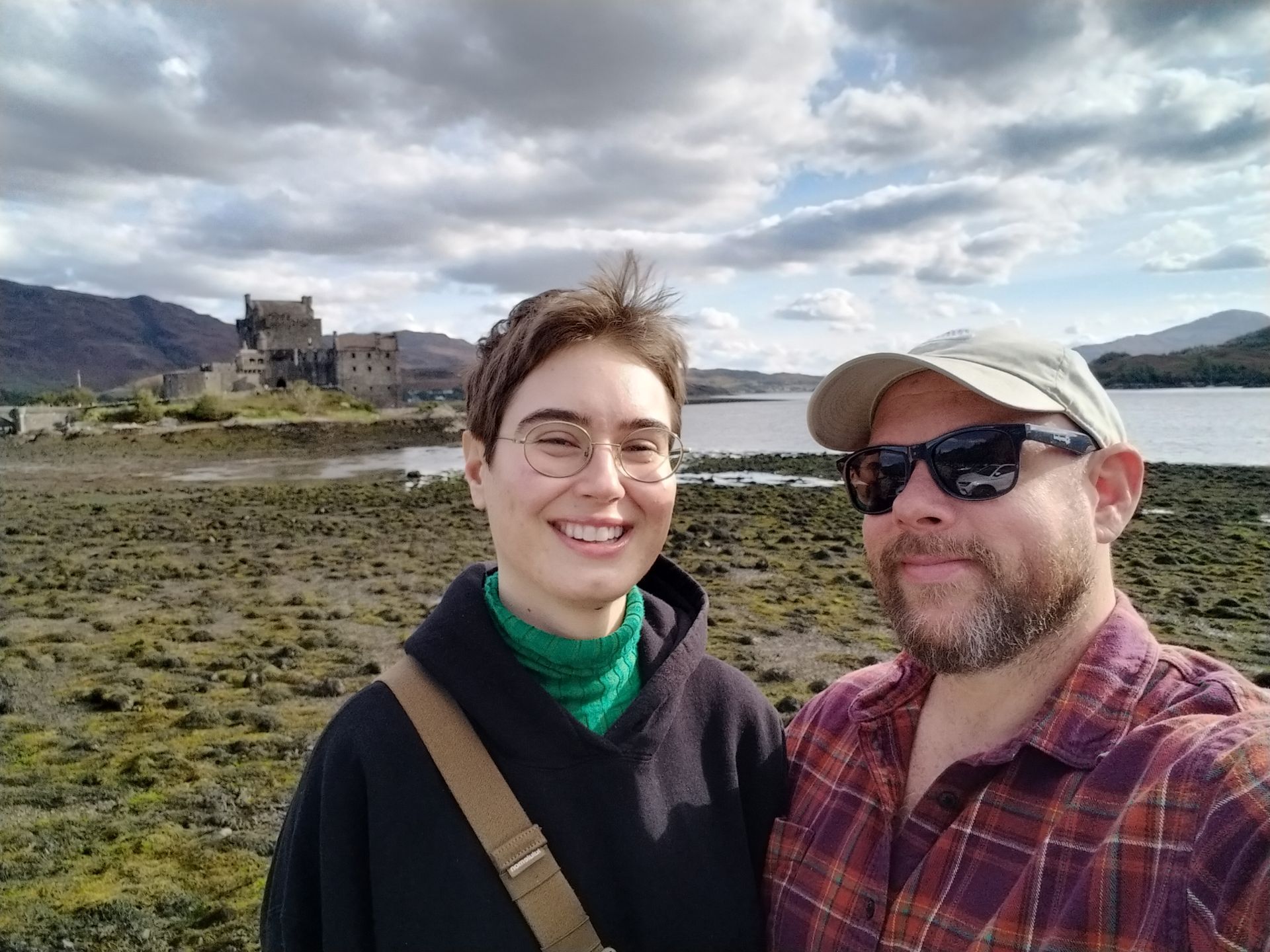 Two people smiling for a selfie in front of a castle by water under cloudy sky.