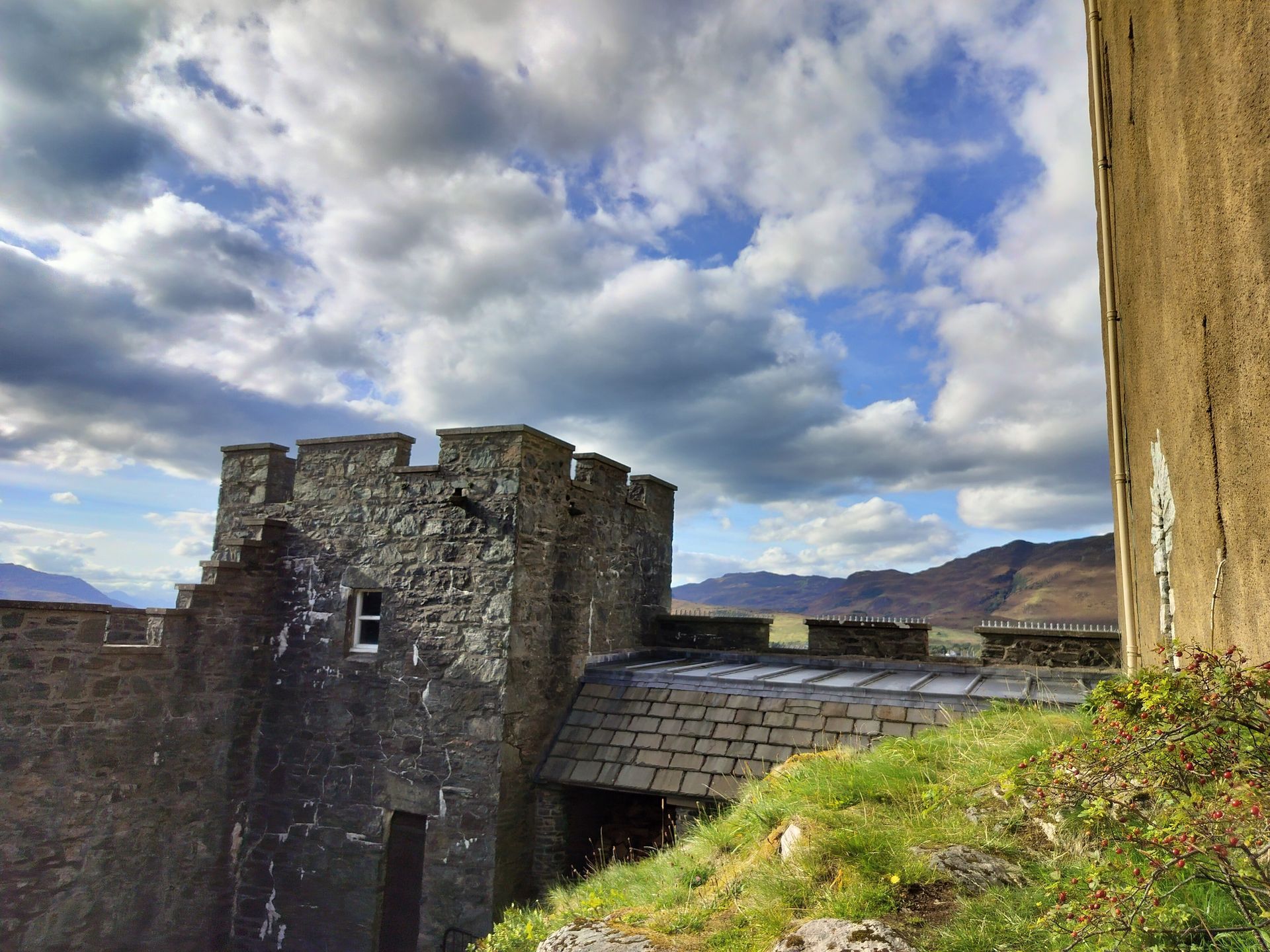 Stone castle turret with crenellations, cloudy blue sky, and mountains in the background.