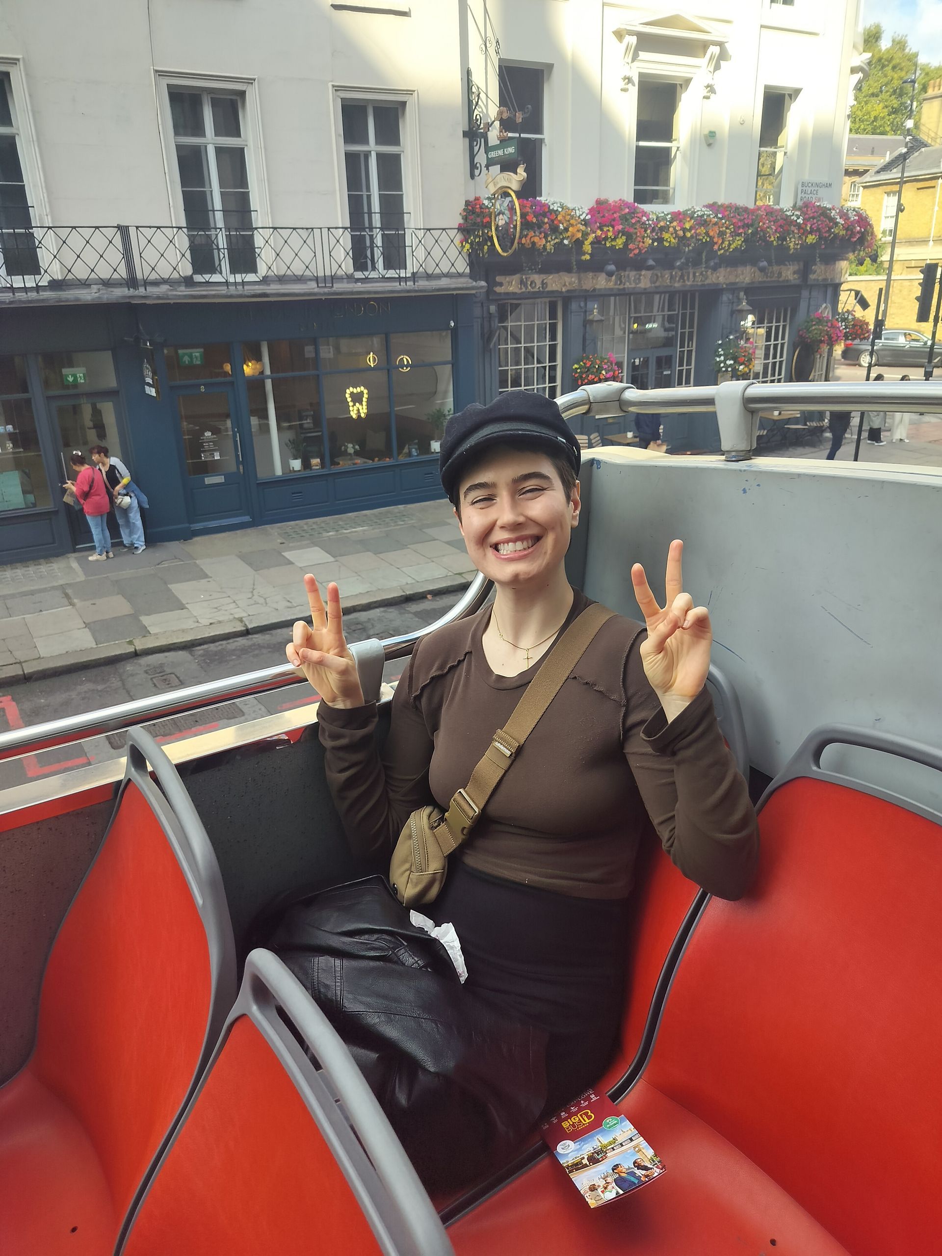 Woman on a red double-decker bus, smiling and giving peace signs. London street scene background.