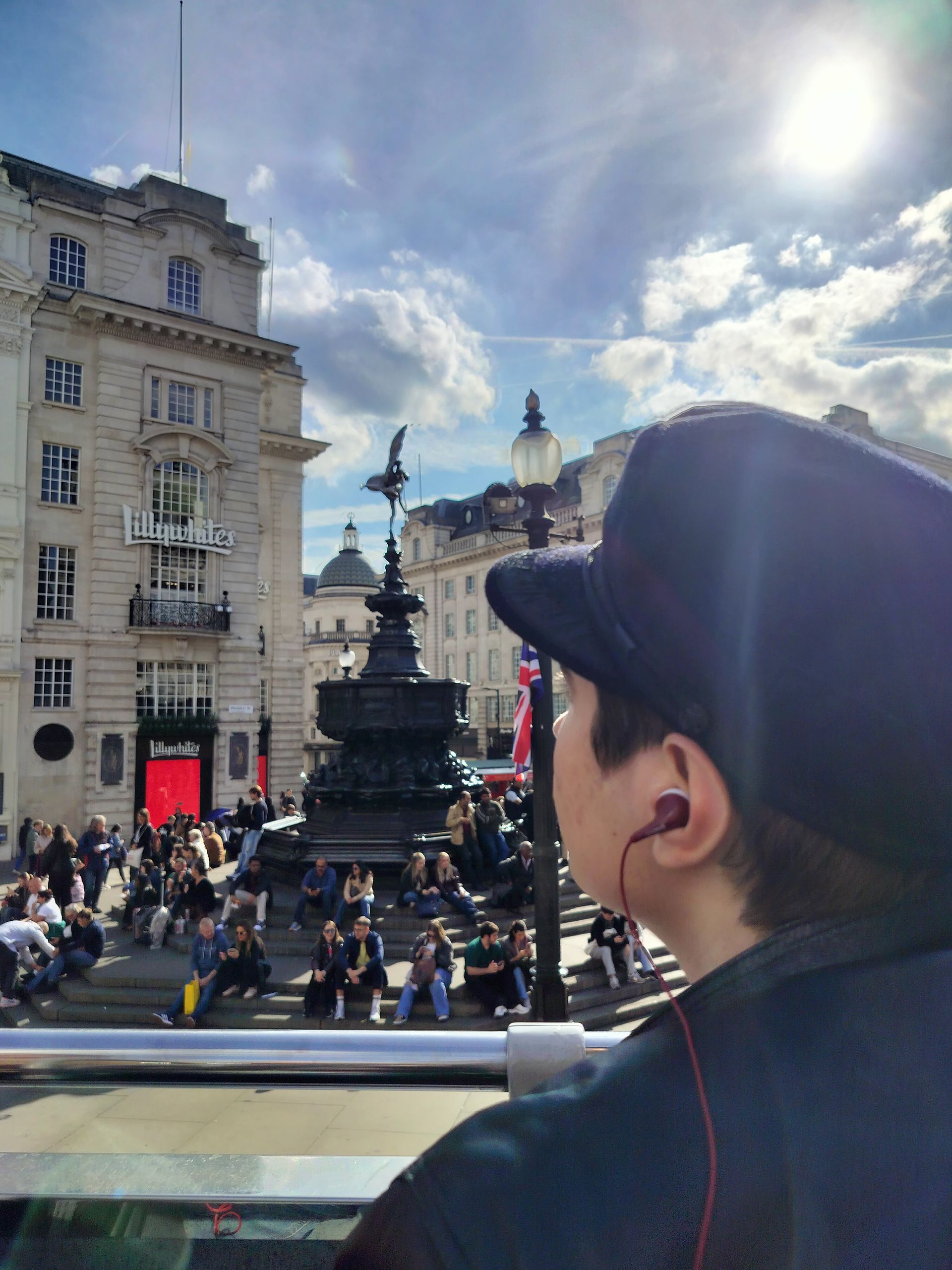 Person wearing a hat looking at Piccadilly Circus, London, statue in center, bright sun, people.