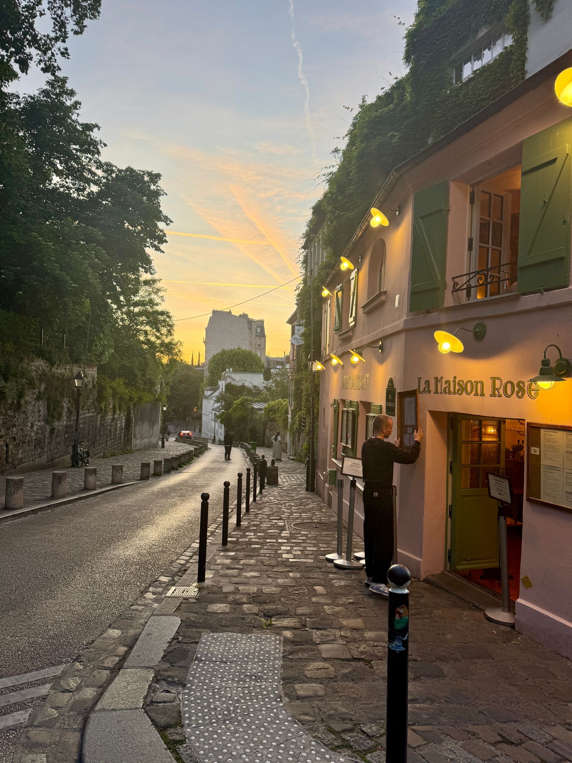 Cobblestone street in Paris with a pink building, person adjusting a sign. Sunset colors in sky.