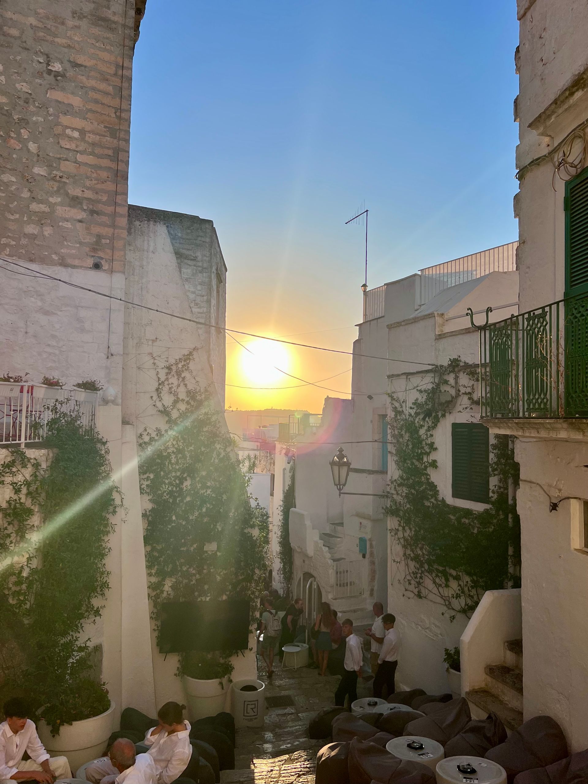 Narrow alley in a whitewashed town, sun setting in the distance. People seated at tables. Green vines on walls.