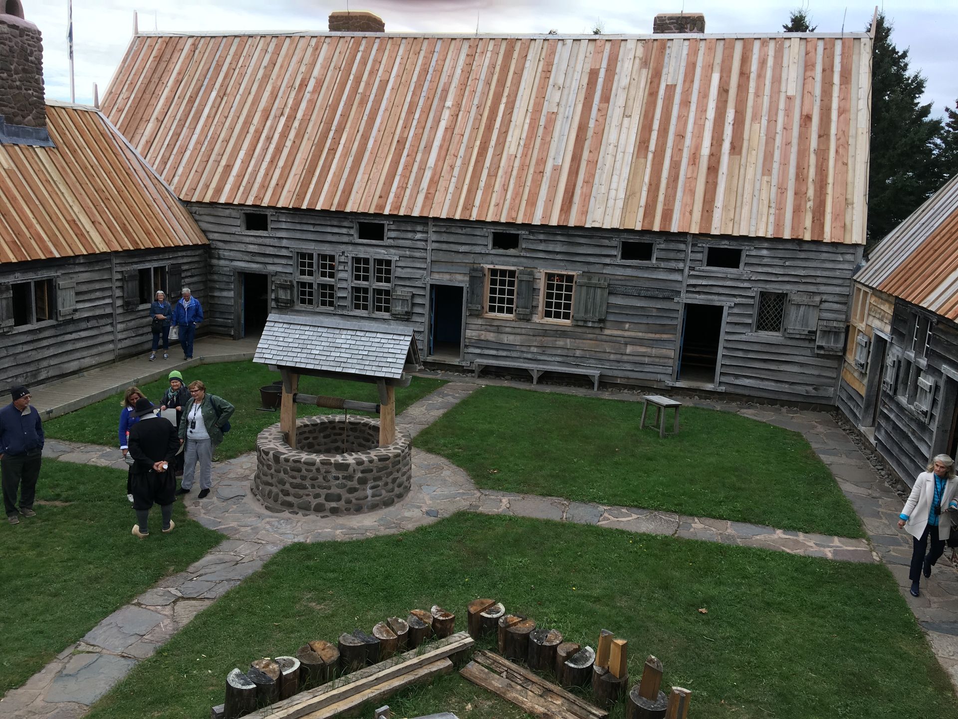 Log buildings with rusty metal roofs surround a central courtyard, people walking around a well.