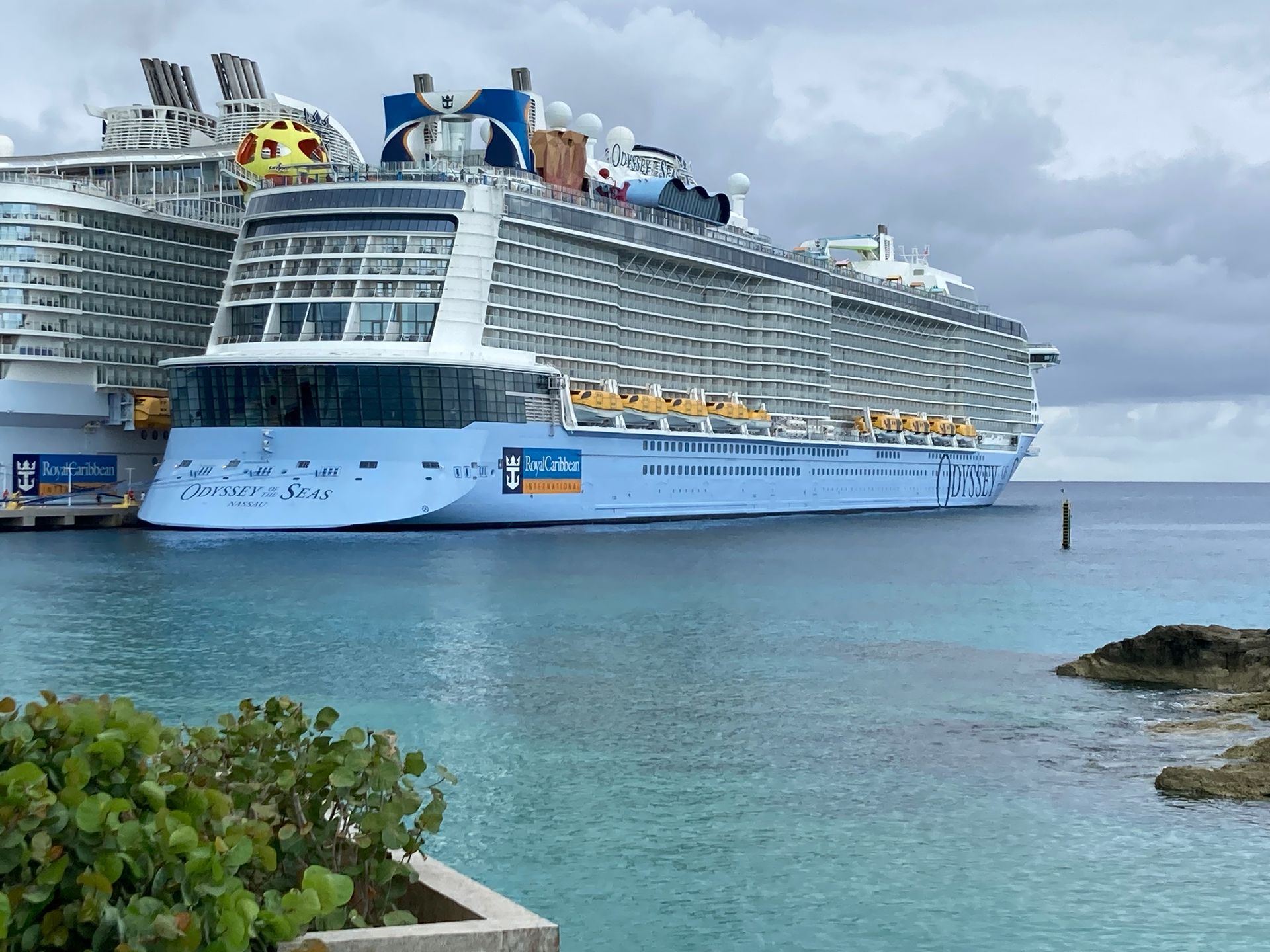 Large blue cruise ship docked at a port, turquoise water, cloudy sky.