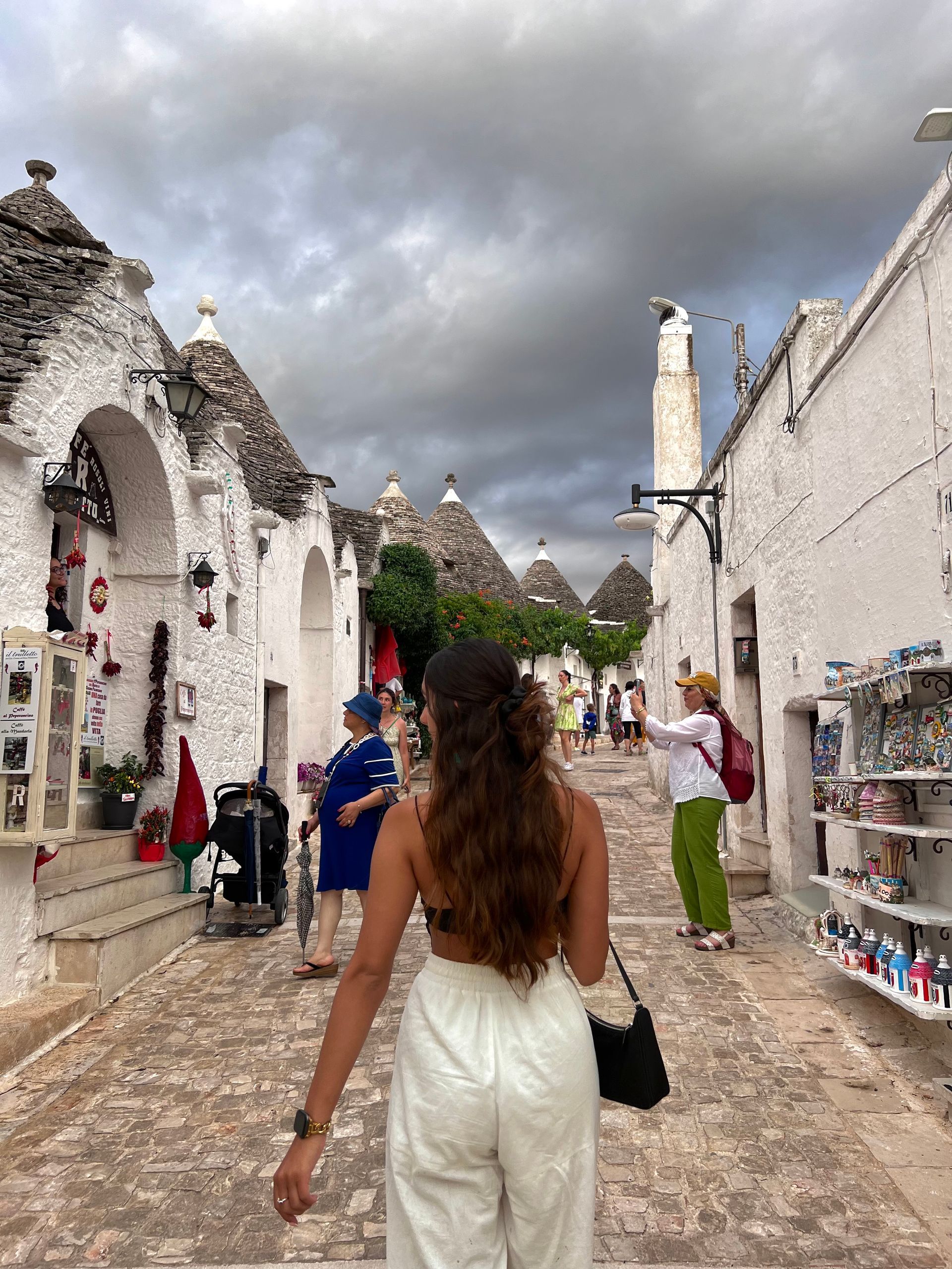 Woman walking cobblestone street lined with white cone-roofed buildings. Cloudy sky overhead.
