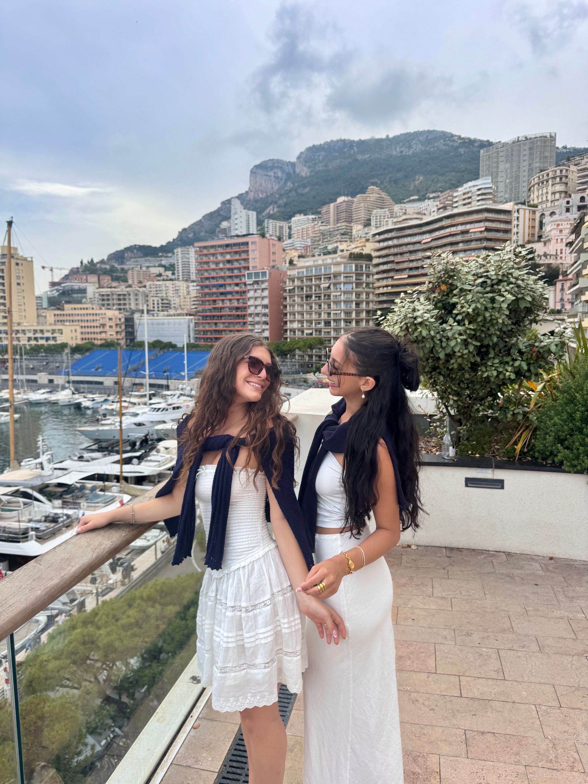 Two women in white outfits, holding hands, smile at each other on a balcony overlooking a marina and city.