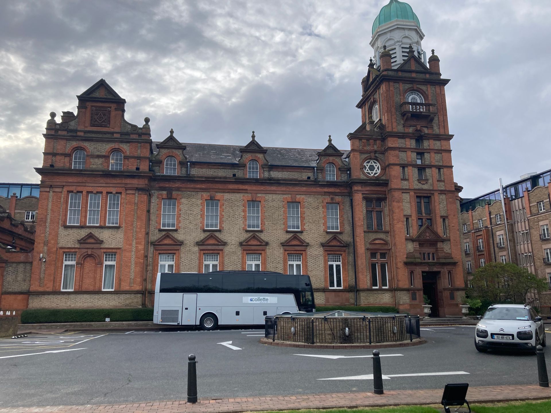 Red brick building with a clock tower and a white bus in front. Cloudy sky.