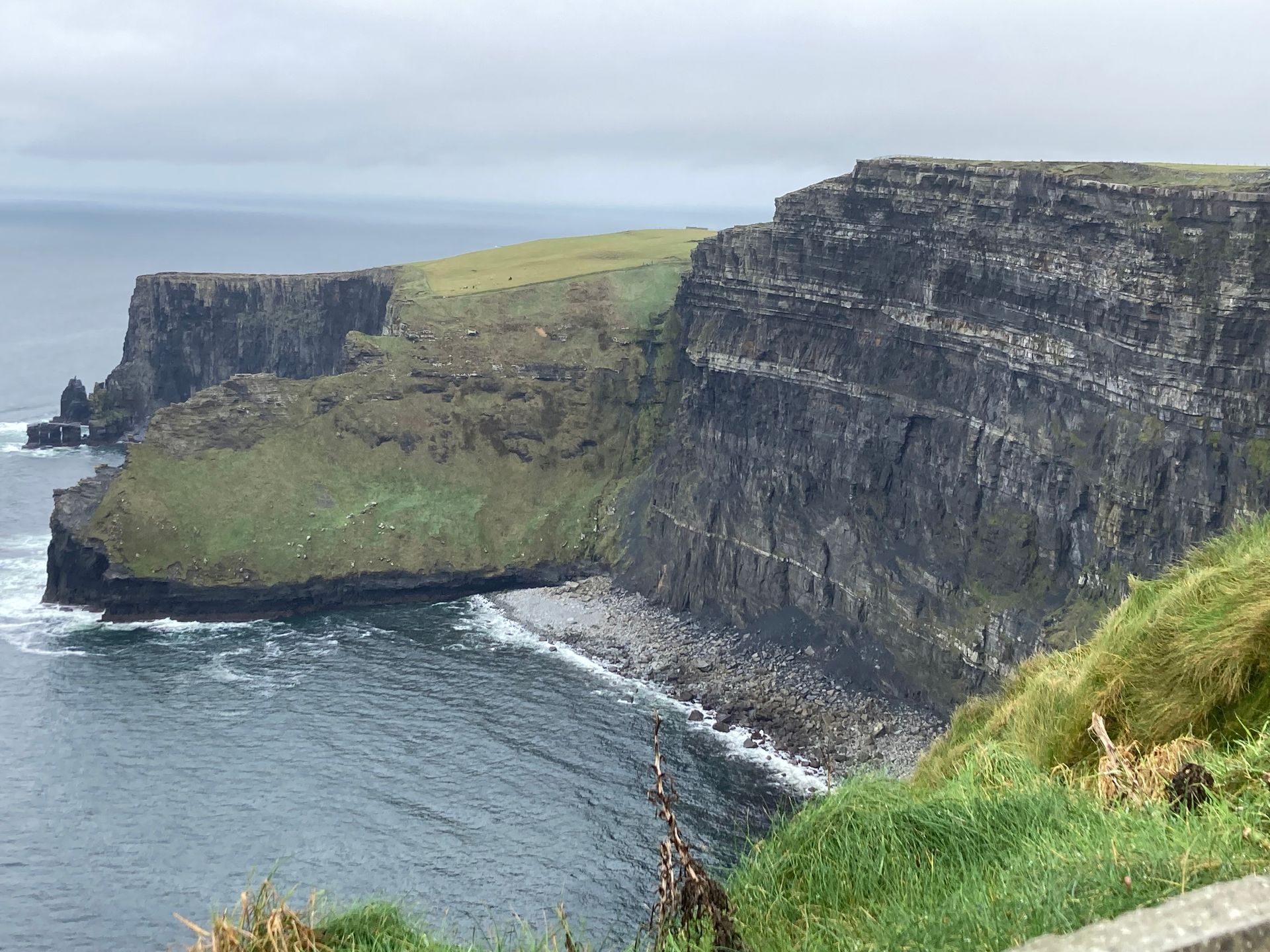Cliffs of Moher, Ireland. Grey, layered rock cliffs meet ocean under overcast sky; green vegetation on top.