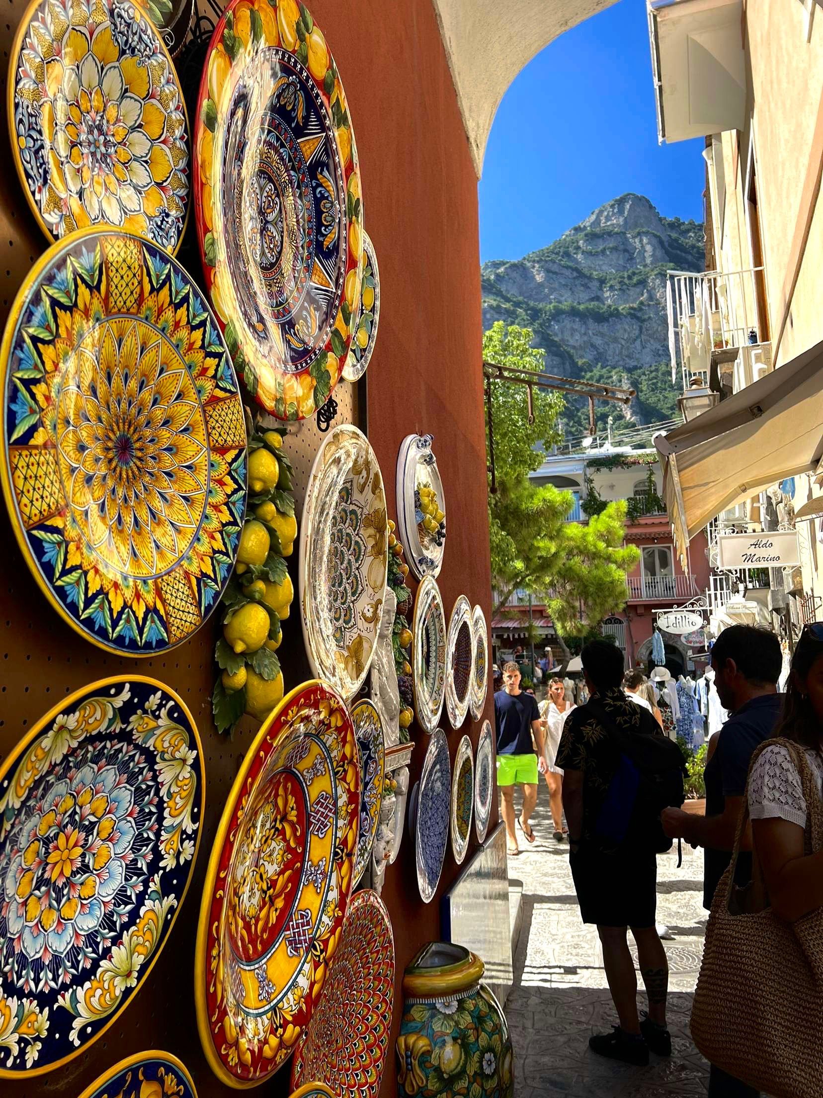 Decorative plates on a wall in Positano, Italy, with a view of the mountains and people walking.