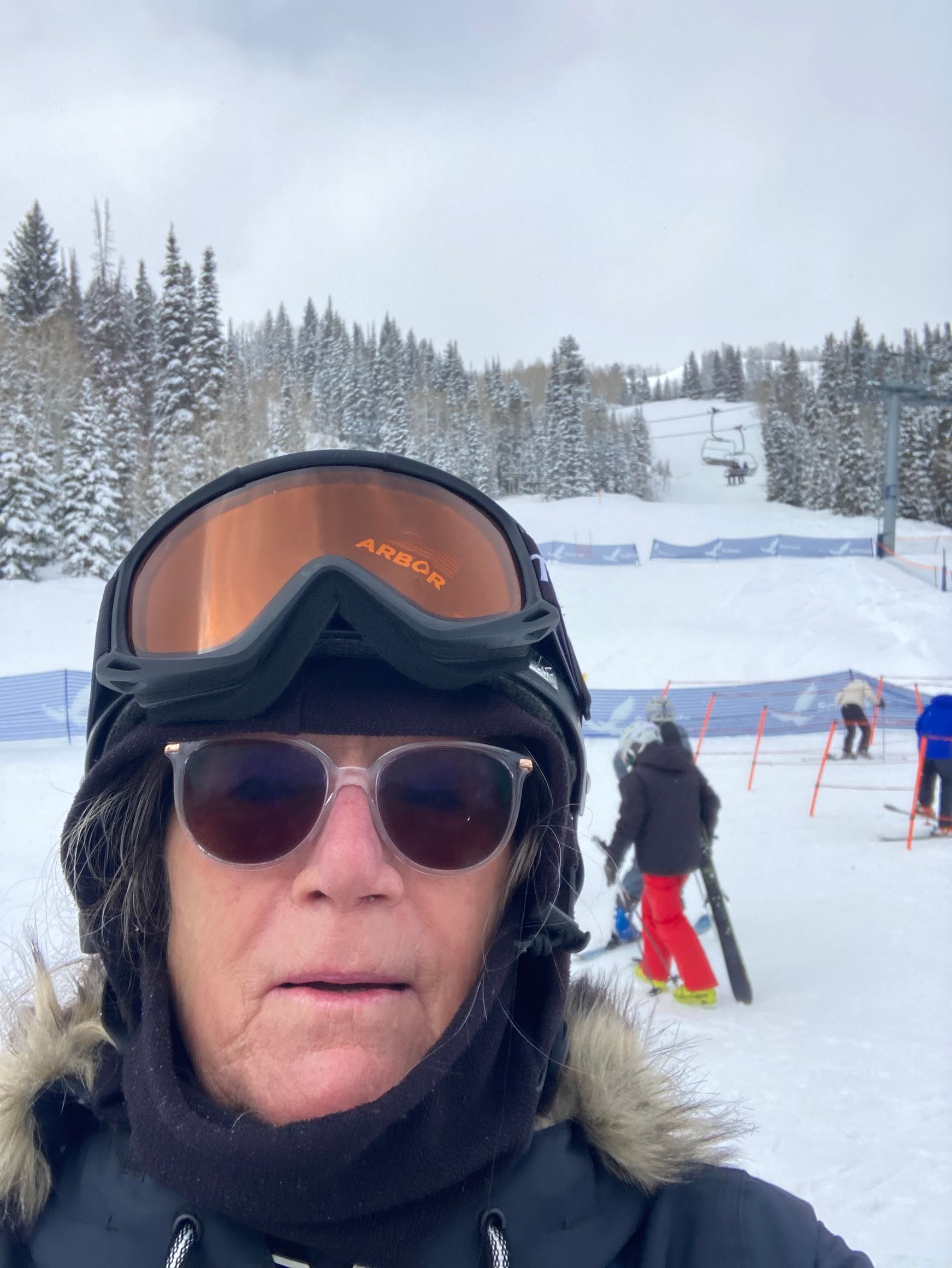 Person in snow gear with goggles and sunglasses, at a ski resort, snow-covered mountain in the background.