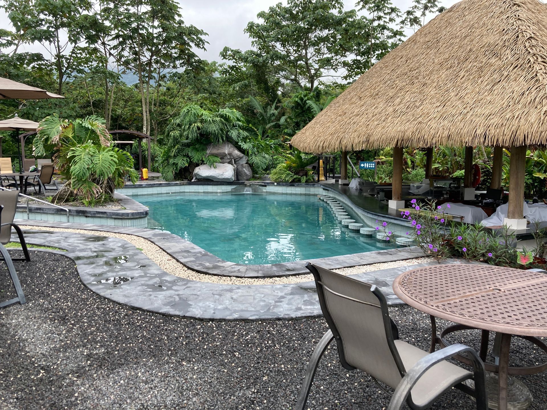 Swimming pool next to a thatched-roof structure, surrounded by tropical plants and seating areas.