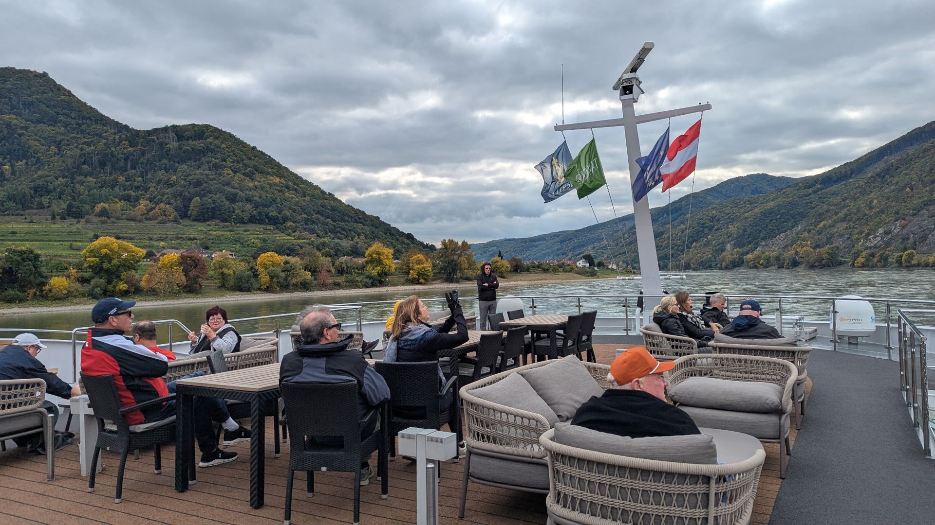 People relaxing on a ship deck with tables and seating, river and mountains in background.
