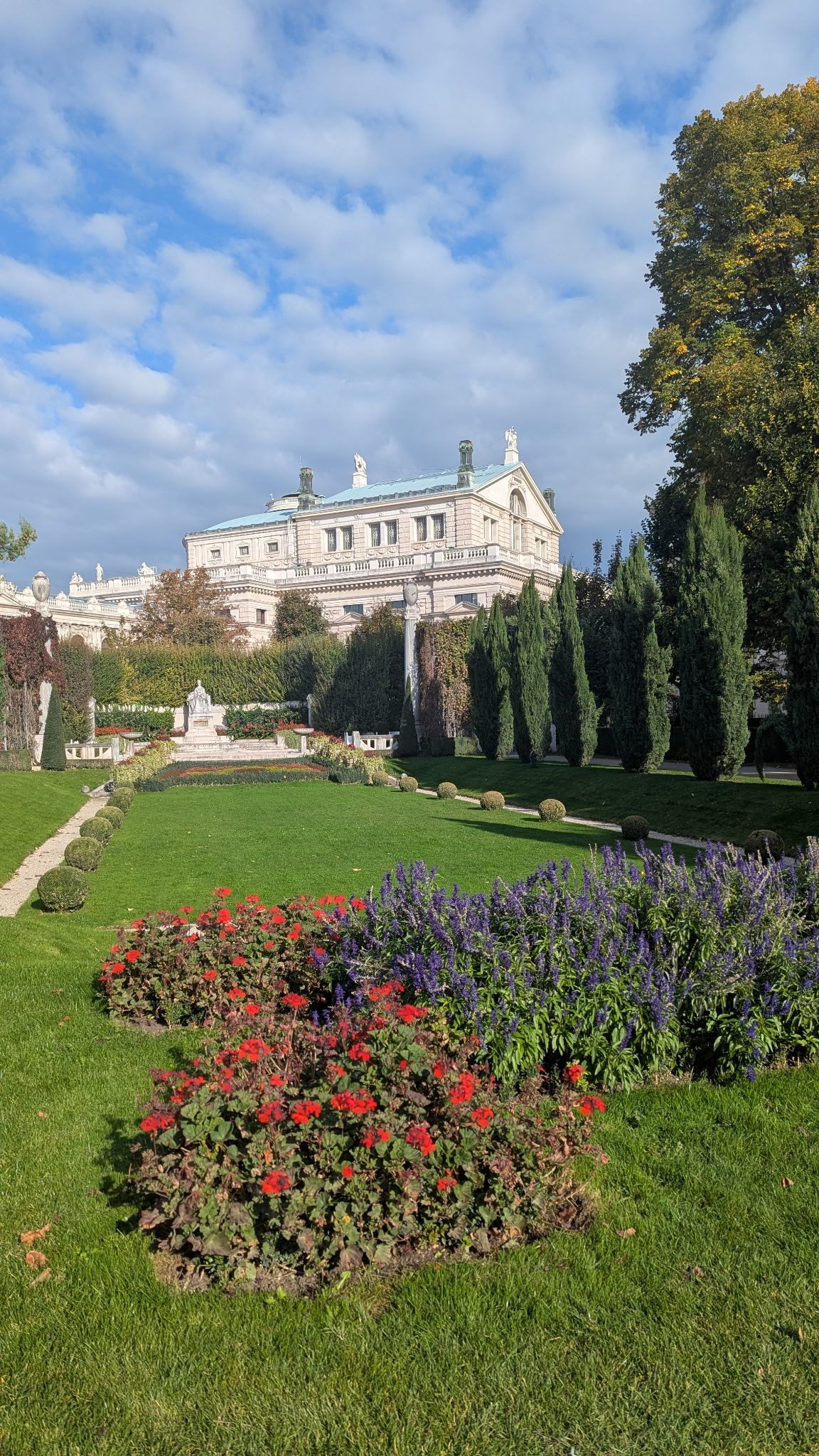 Formal garden with colorful flowerbeds, green grass, and a white ornate building under a cloudy sky.