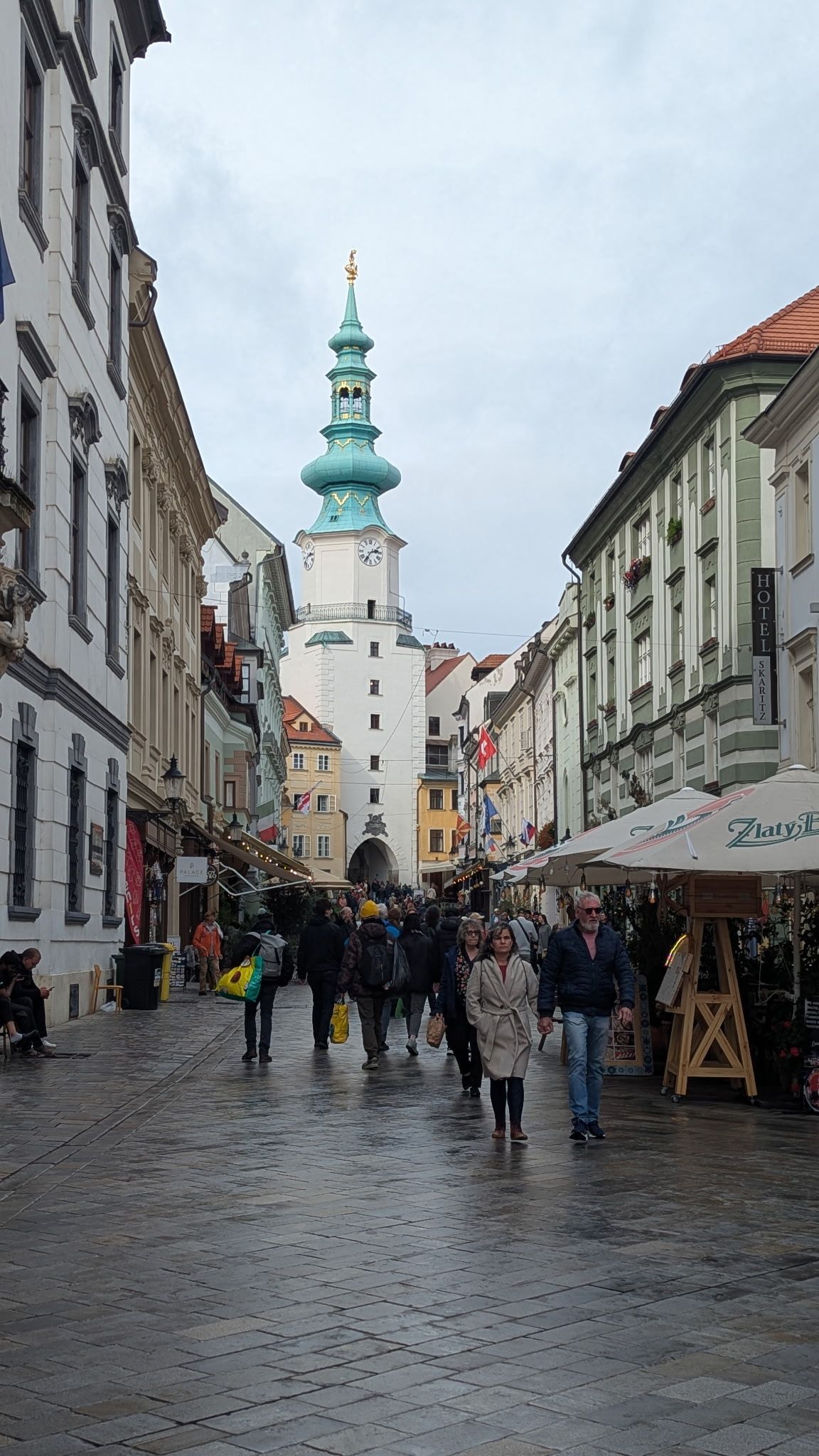 Cobblestone street lined with buildings leads to a tall tower with a green spire, pedestrians stroll, cloudy day.